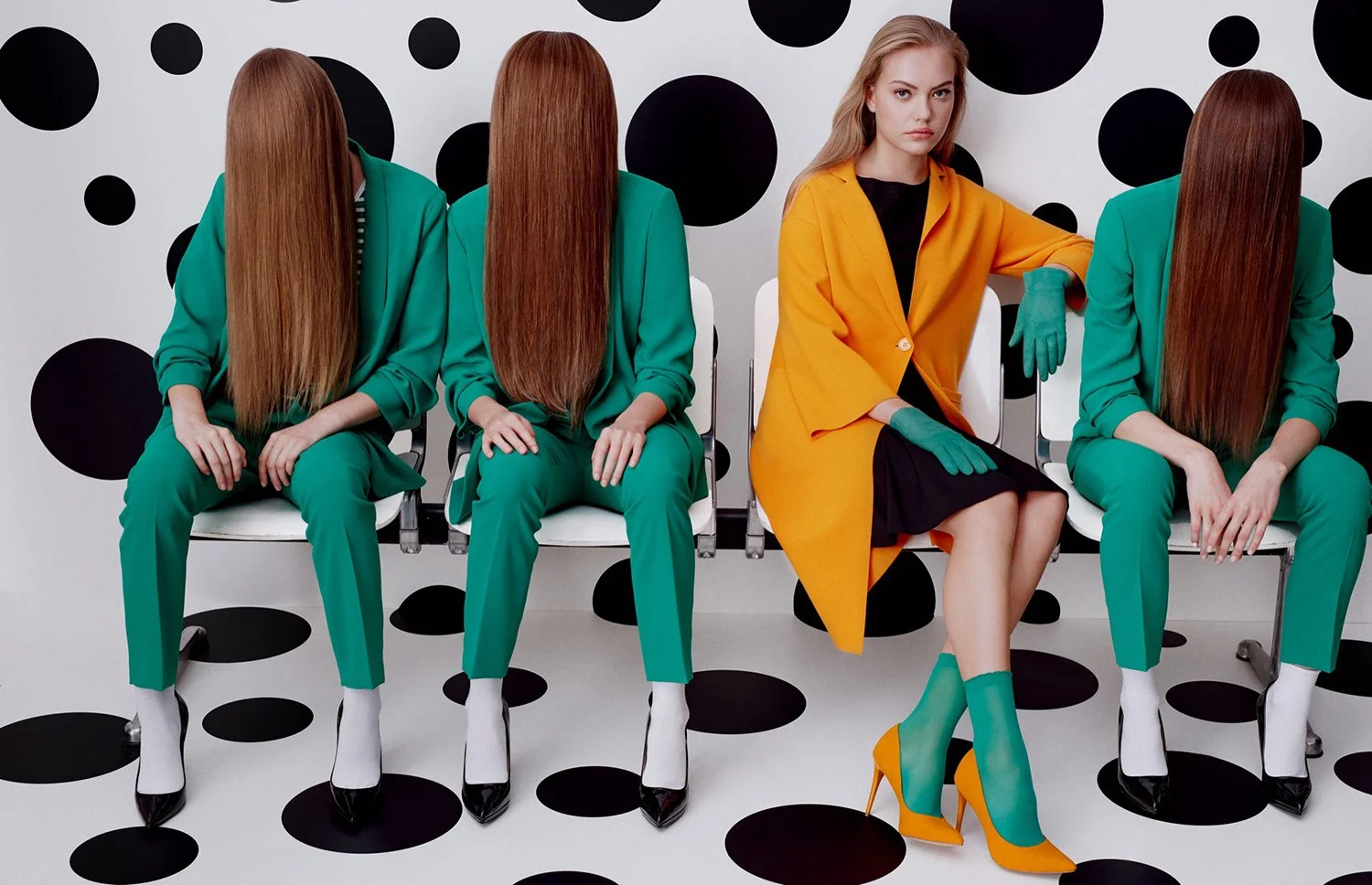 Four women sitting on chairs in front of a black and white polka dot patterned wall. The women on the ends are dressed in green suits with long, straight, reddish-brown hair covering their faces. The second woman from the right wears an orange coat, 