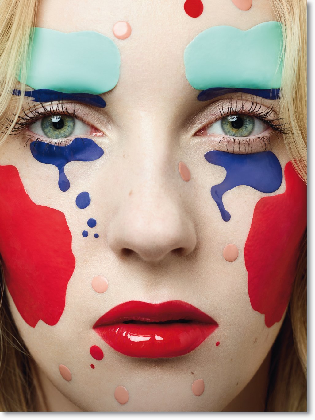 Nahaufnahme eines Gesichts mit bunter Make-up-Kunst, farbigen Akzenten um die Augen, auf den Wangen und am Kinn, rote und blaue Farben, rote Lippen, blasse Haut.