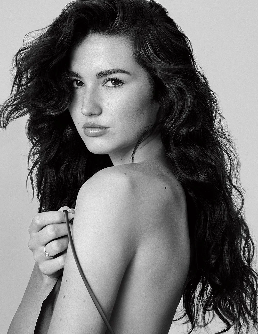 Schwarz-weiß-Fotografie einer jungen Frau mit langen, lockigen Haaren und natürlichem Make-up, die direkt in die Kamera schaut.