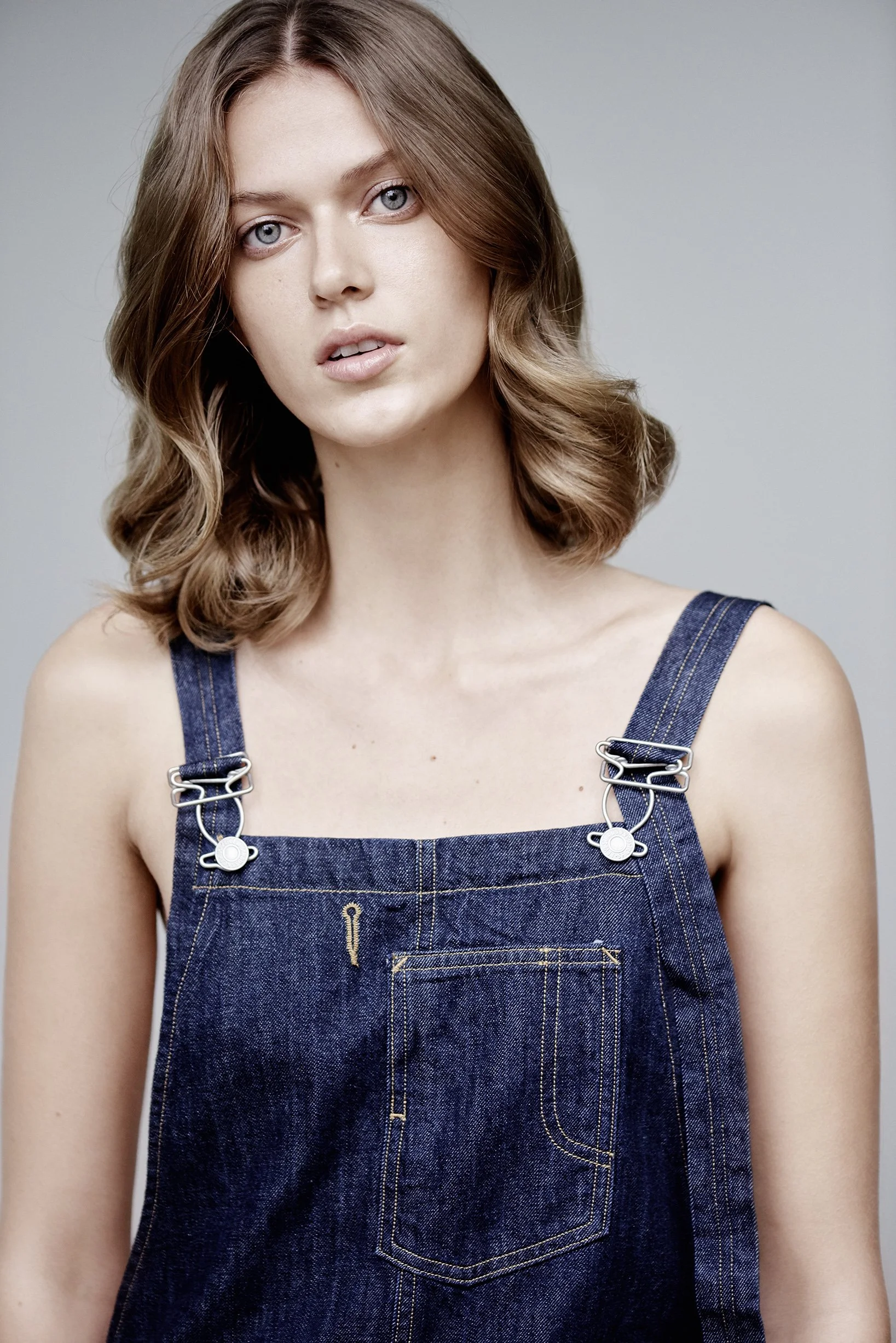 Junge Frau mit braunen, gewellten Haaren trägt dunkelblaue Jeans-Shorts, blickt direkt in die Kamera, neutraler Hintergrund.