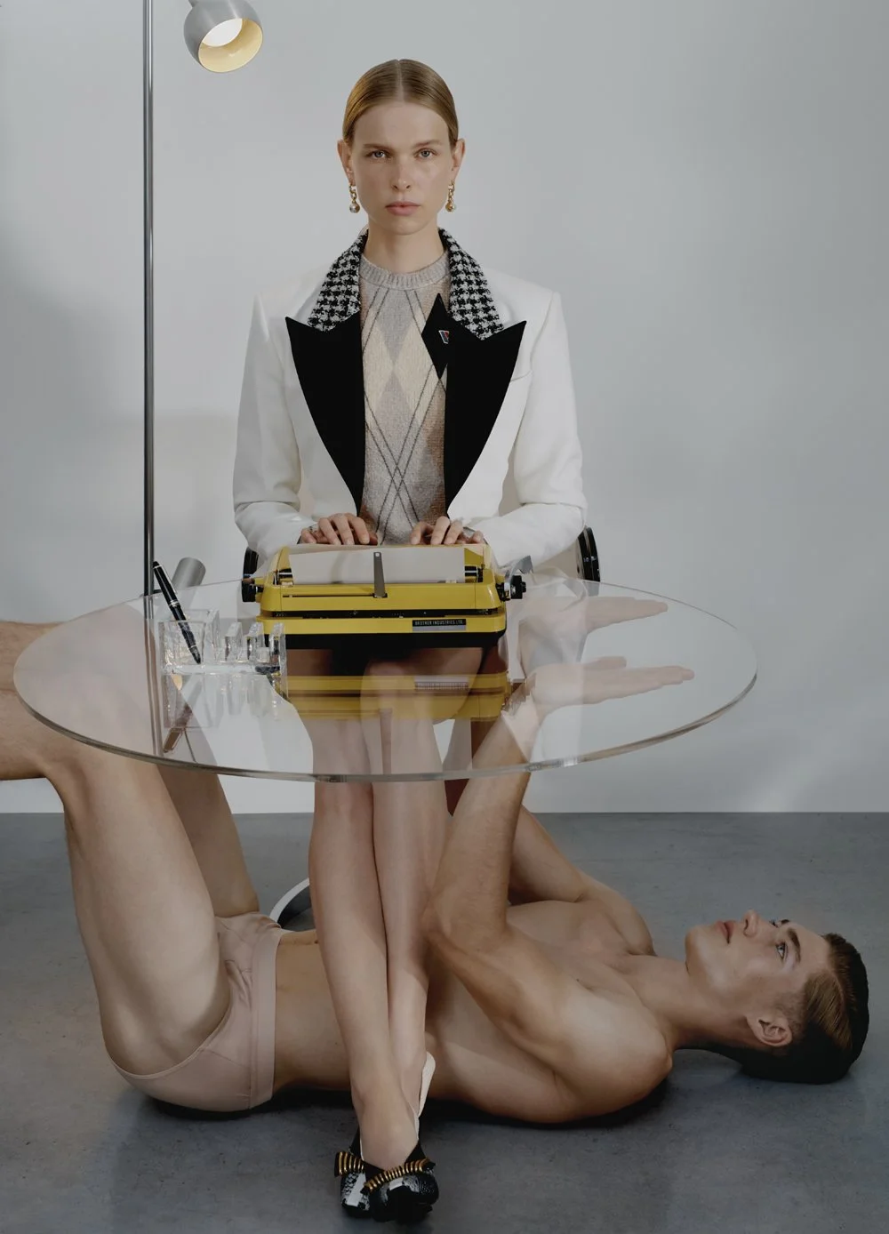 Frau im eleganten Outfit sitzt hinter einem runden Glas-Desk, während ein nackter Mann unter dem Tisch liegt und seine Beine um die Beine der Frau schlingt.