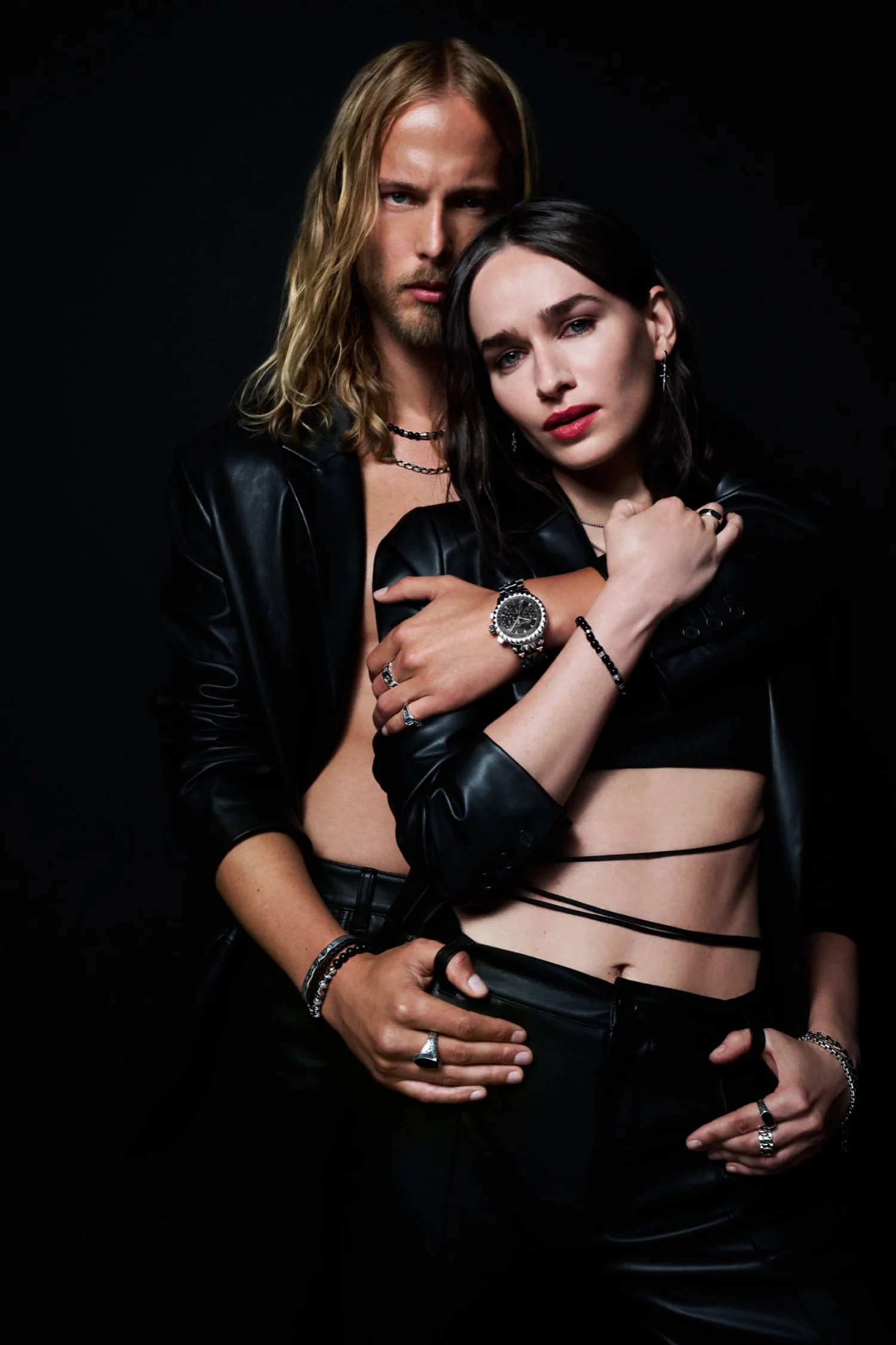 A man and woman pose together against a black background, both dressed in black leather outfits with jewelry, looking directly at the camera.