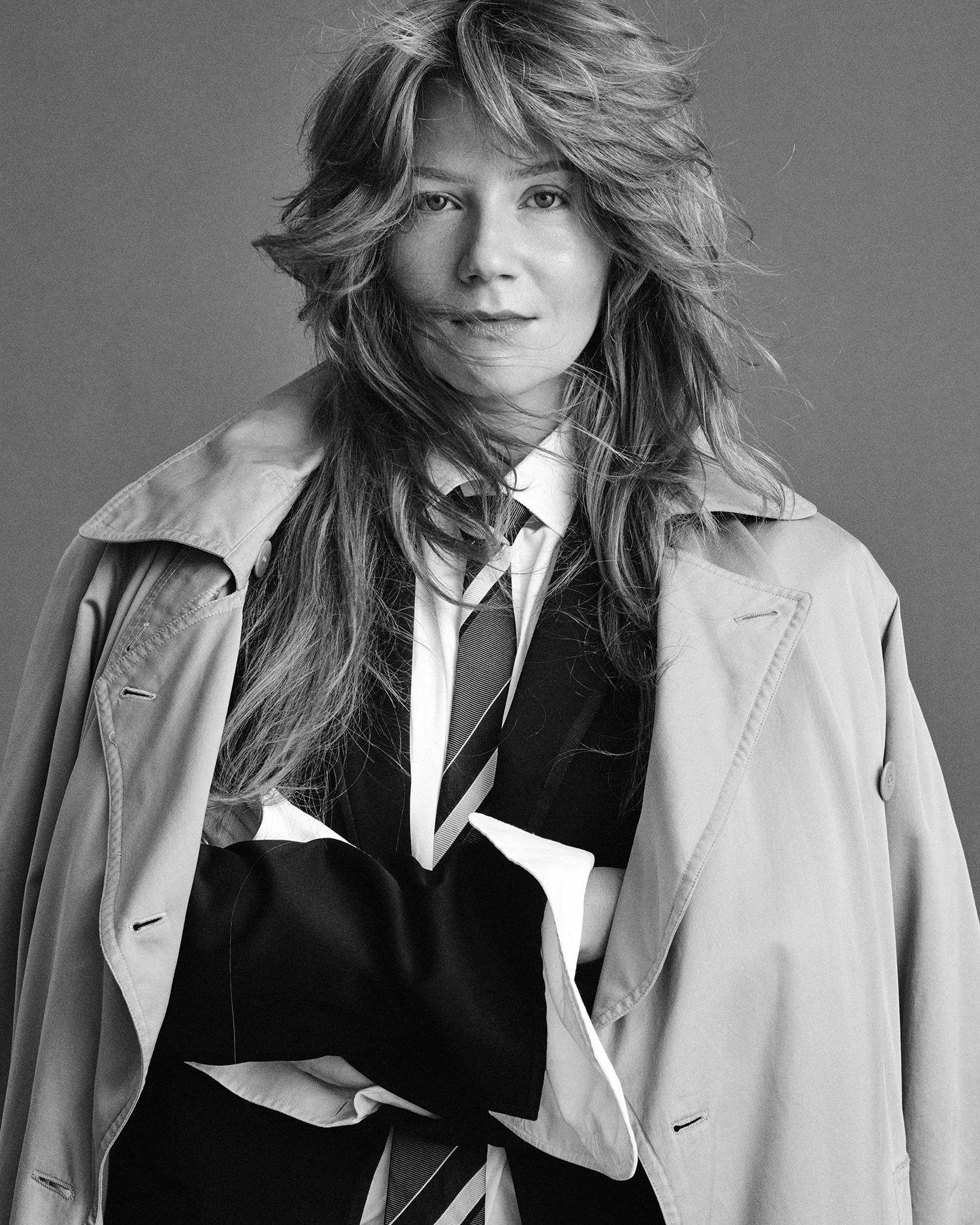 Black and white portrait of a woman with wavy hair wearing a trench coat, shirt, and tie.
