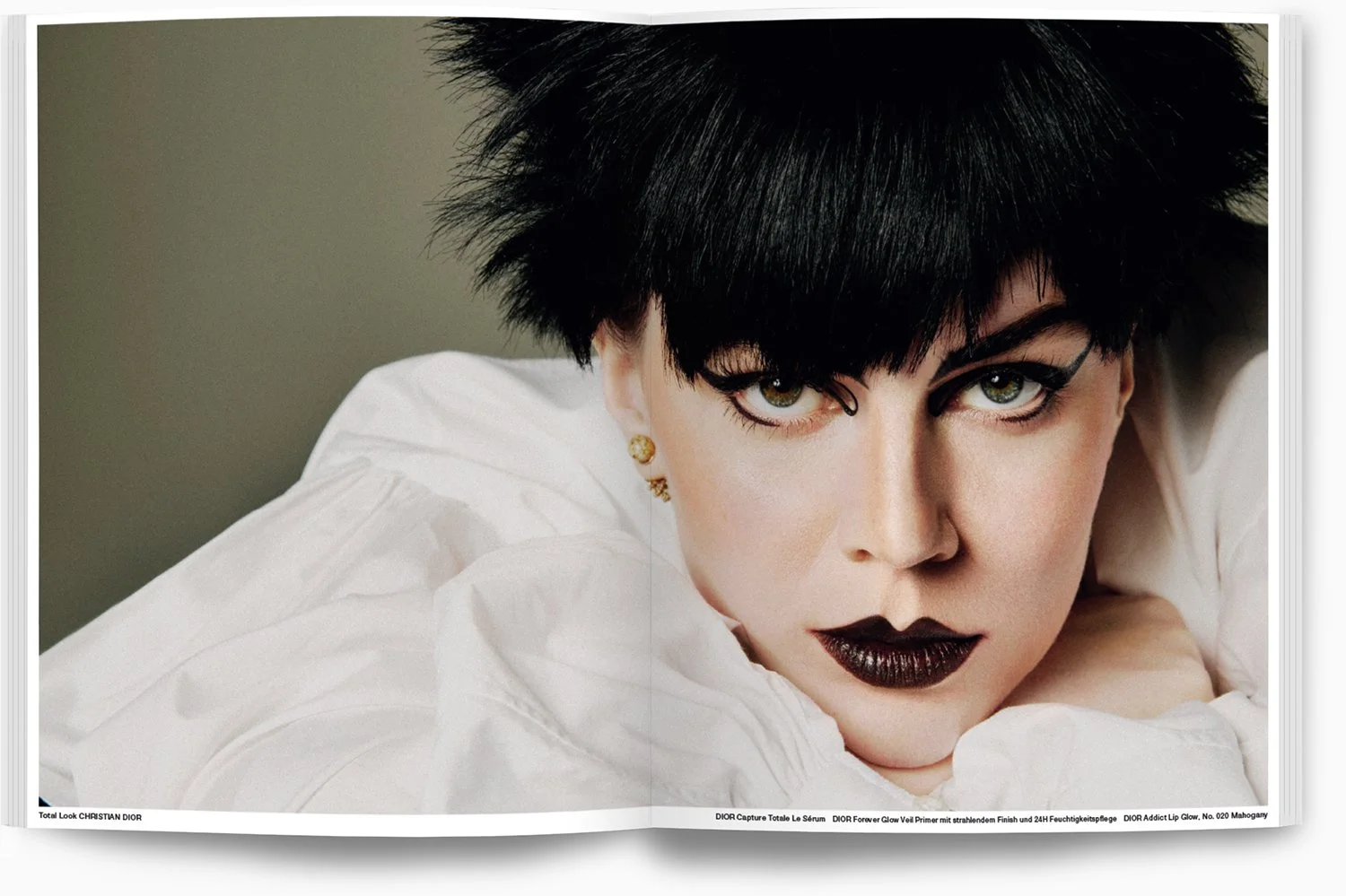 Nahaufnahme eines Models mit dunklem, wild gestyltem Haar, silbernen Ohrringen, dunklem Lippenstift, weißen Kleidung und intensiven Blick, liegend auf einem weißen Kissen.