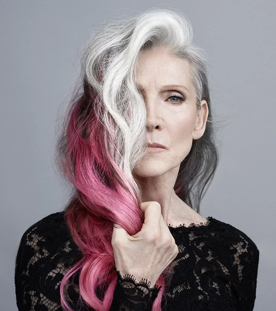 Porträt einer älteren Frau mit silbergrau-weißen Haaren, teilweise gefärbt in Pink, trägt schwarze Spitze-Bluse, vor grauem Hintergrund.