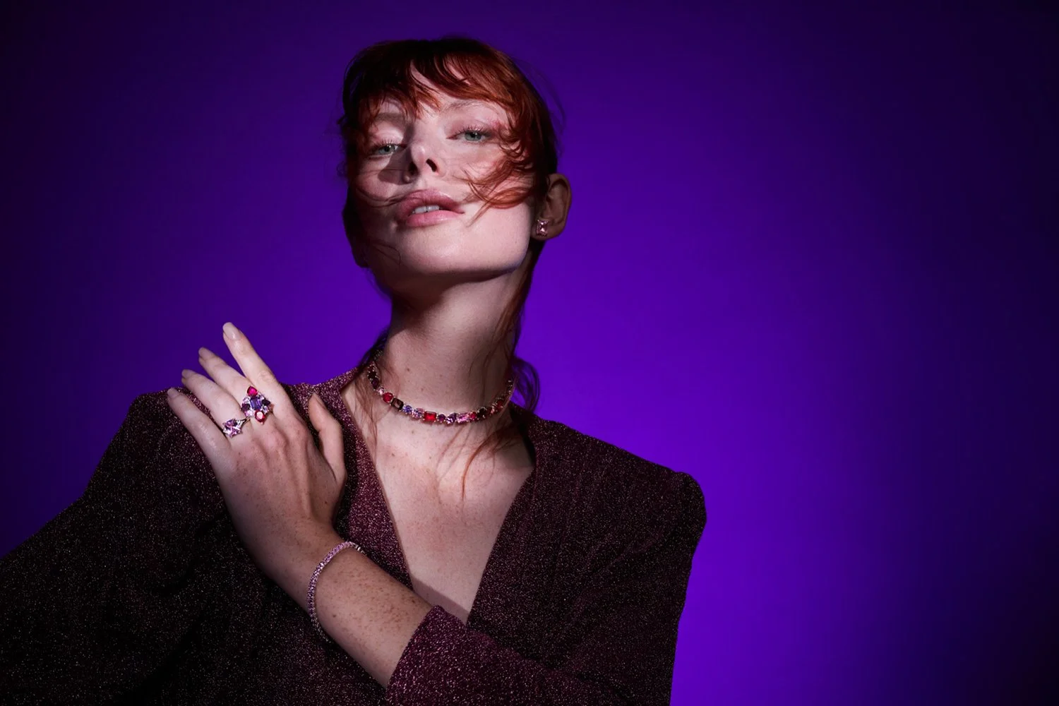 A woman with red hair, wearing jewelry including rings, a necklace, and earrings, posing against a purple background.