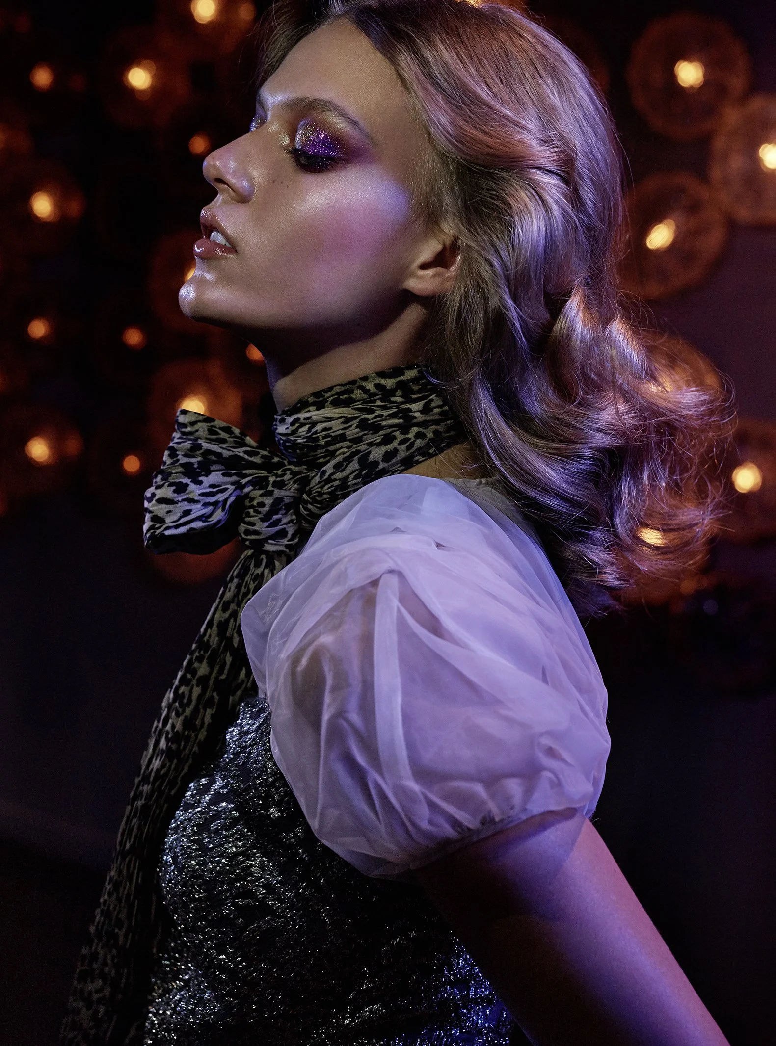 Eine Frau mit auffälligem Make-up, silbrigen Locken, im Profil auf Dunklem Hintergrund mit leuchtenden, orangefarbenen Lichtern im Hintergrund.