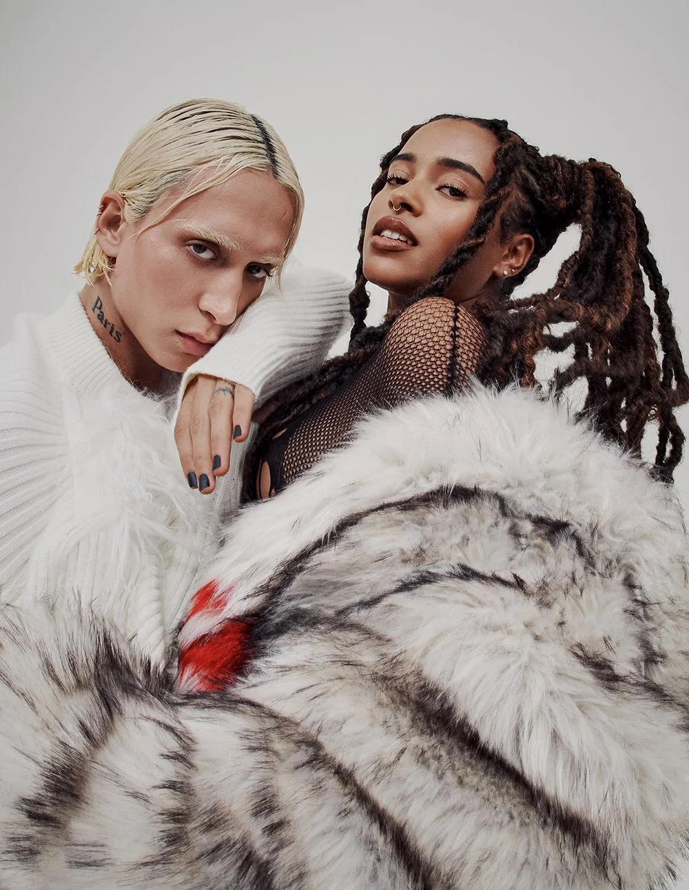 Zwei junge Frauen posieren gemeinsam vor einem neutralen Hintergrund, eine mit blonden Haaren, die andere mit dreadlocks, beide tragen modische Kleidung.