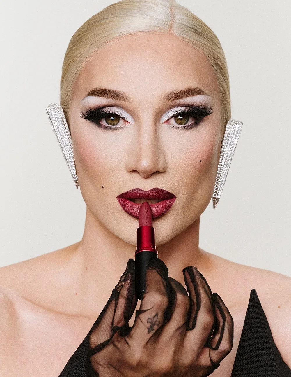 Eine Frau mit blonden Haaren, auffälligem Make-up, großen Ohrringen, hält einen Lippenstift vor den Lippen und trägt einen schwarzen, durchsichtigen Handschuh.