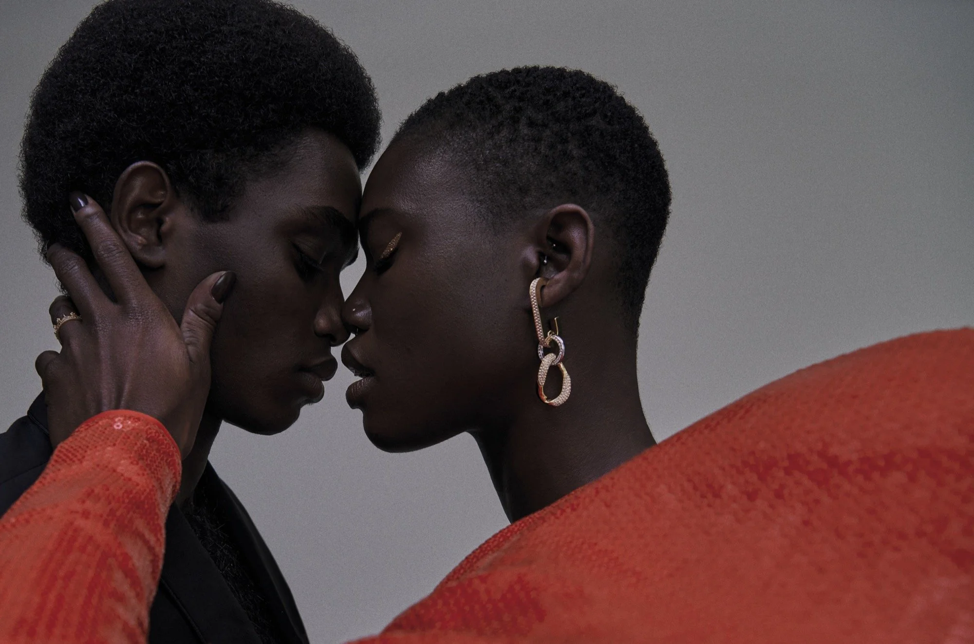 Zwei schwarze Frauen stehen sich eng gegenüber, ihre Stirn berühren sich. Eine trägt eine orange Kleidung, die andere dunkle Kleidung. Sie haben die Augen geschlossen, der Moment wirkt intimer und emotional.