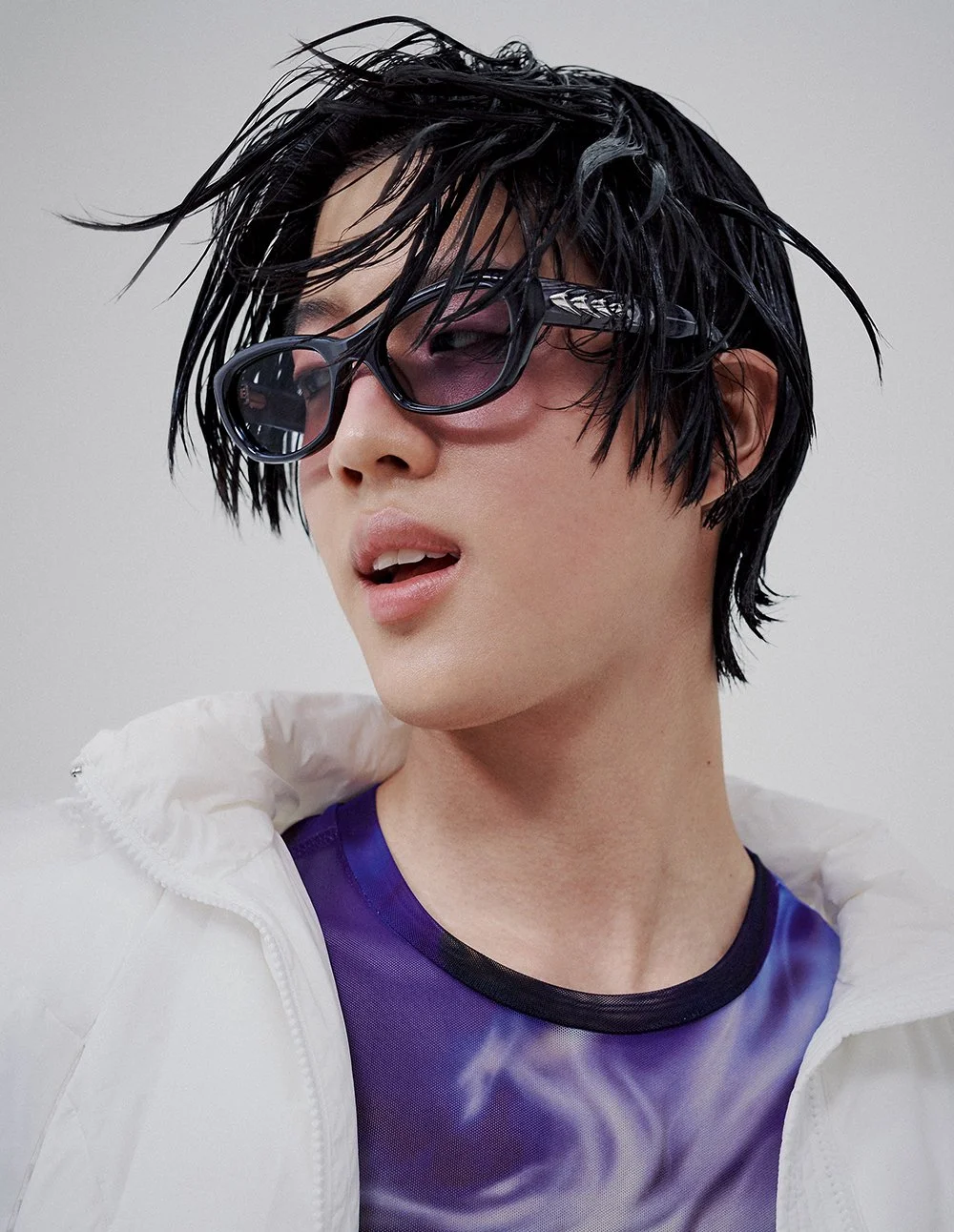 Junge Person mit nassem, schwarzem, strukturiertem Haar trägt schwarze Sonnenbrille mit Musterrahmen, lila-türkisfarbenem Oberteil mit Rauchmuster und einer weißen Jacke, vor grauem Hintergrund.