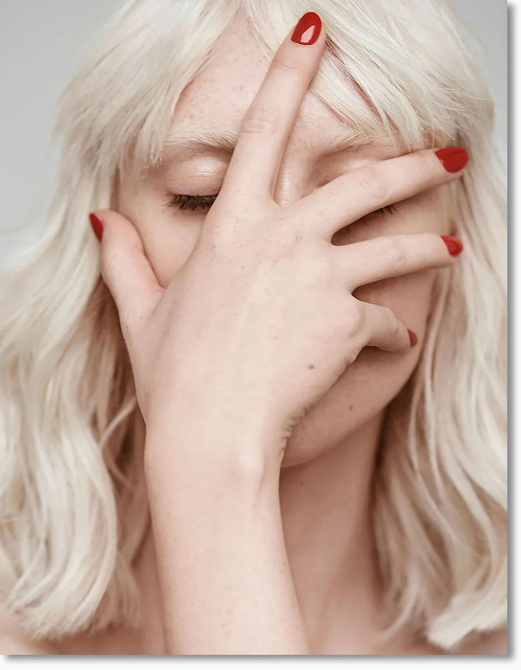 Nahaufnahme eines Frauenkopfes mit blondem, lockigem Haar, die Augen sind geschlossen, eine Hand bedeckt das Gesicht mit roten Nagellack auf den Fingern.