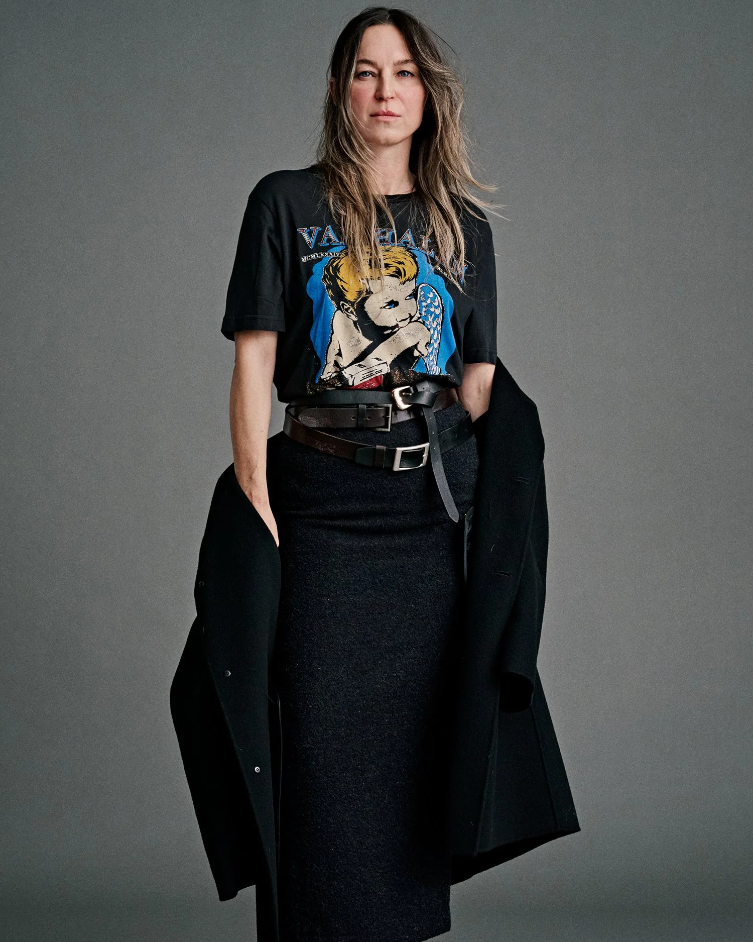 Woman in black graphic t-shirt, black skirt, and black coat holding coat in hand, standing against gray background.