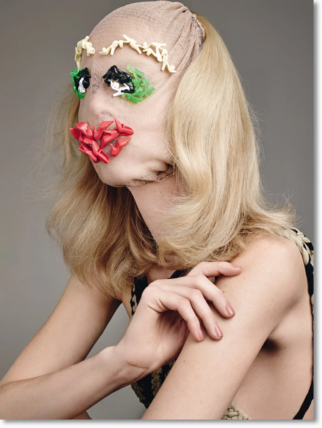 Verhülltes Gesicht einer Frau mit einer Maske, die aus bunten Klammern besteht. Sie hat blonde, gewellte Haare und trägt ein schwarzes Kleid mit Tierenprint.