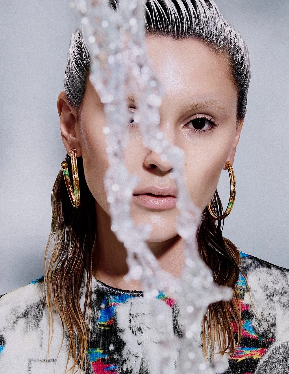 Nahaufnahme eines Gesichts einer jungen Frau mit nassen, braunen Haaren, die durch eine schmale, unregelmäßige Wasserlinie halb verdeckt ist, während ihre rechten Ohrringe große goldene Creolen sind und sie ein bunt gemustertes Oberteil trägt.