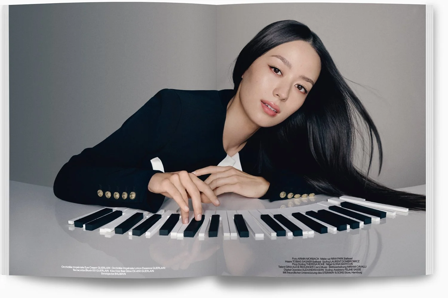 Junge Frau mit schwarzen Haaren liegt auf einem Klavier, Kopf auf die Arme gestützt, Blick in die Kamera, schwarzes Jackett mit goldenen Knöpfen, grauer Hintergrund.