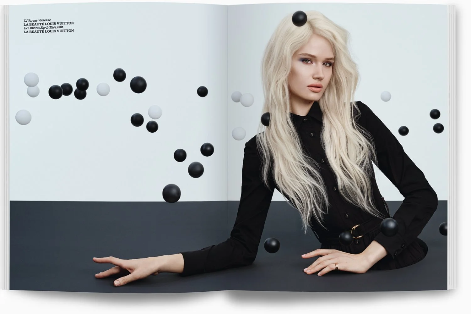 Eine Frau mit langen, lockigen, blonden Haaren trägt ein schwarzes Kleid. Sie liegt auf einem schwarzen Tisch, umgeben von schwebenden schwarzen und weißen Kugeln, vor einem hellen Hintergrund.