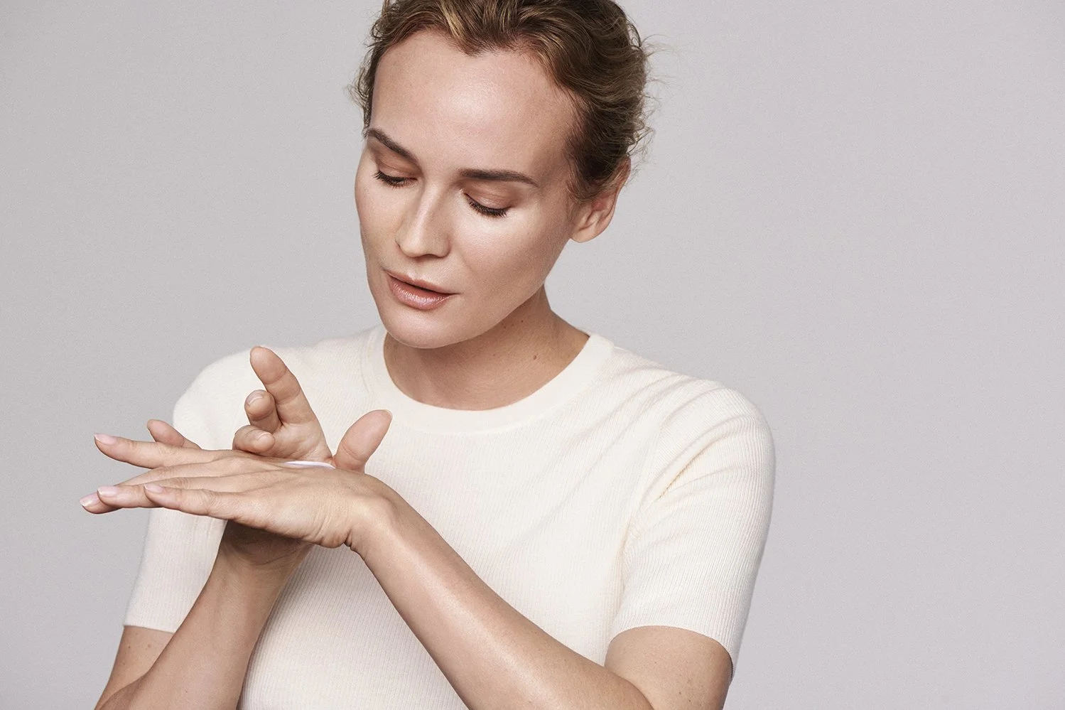 A woman with short, styled hair applying cream or lotion to her hands against a plain, light background.