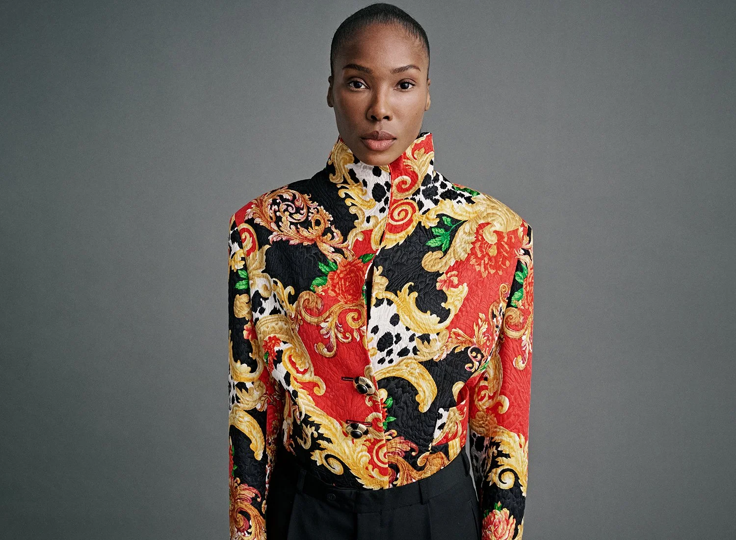 A woman with a high ponytail wearing a colorful, ornate patterned jacket with gold, red, black, white, and green designs against a plain gray background.