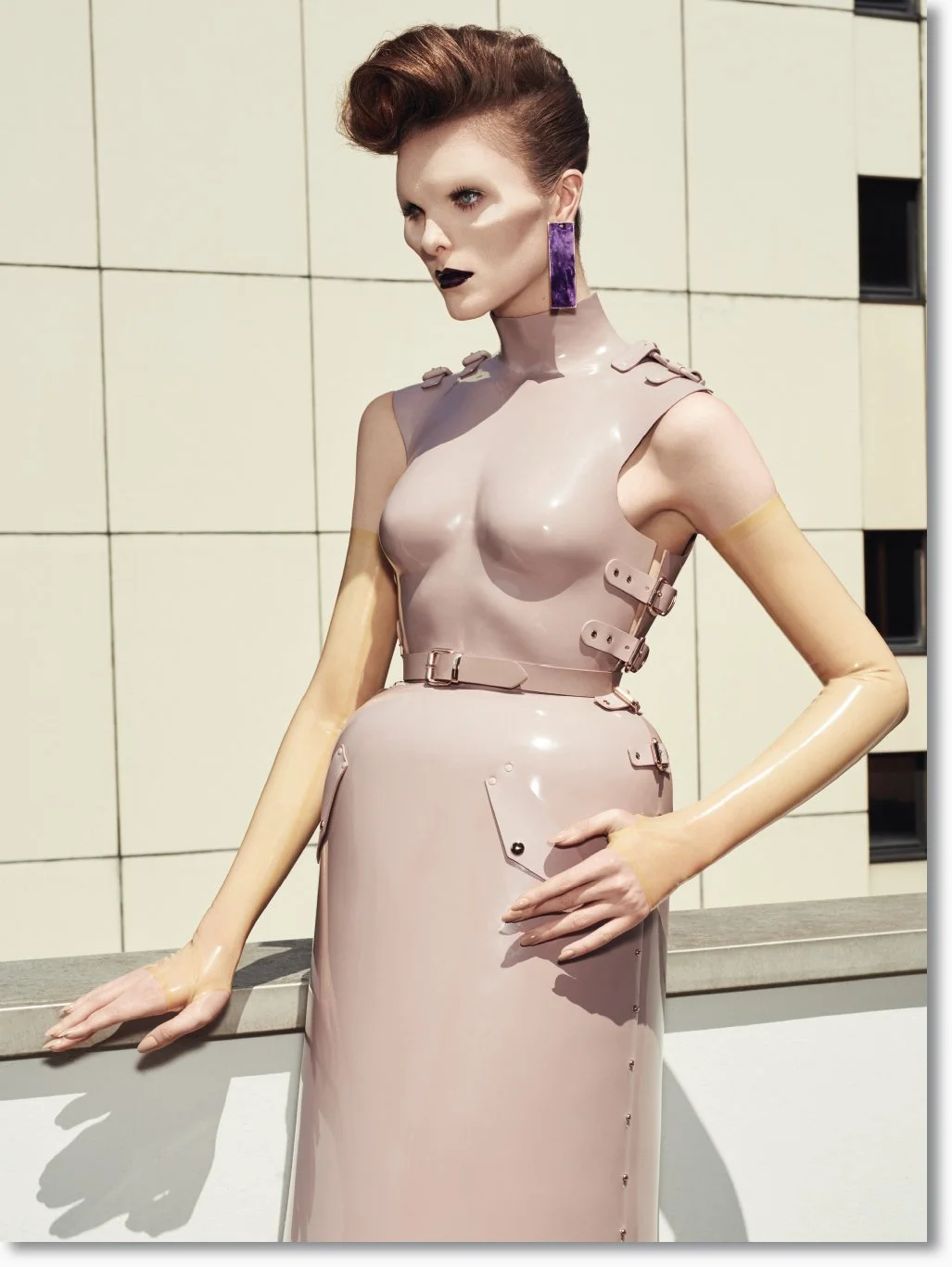 Eine Modenschau-Braut trägt ein futuristisches Kleid aus glänzendem, beige-rosafarbenem Material mit Schnallen und Verzierungen, ergänzt durch auffällige, große lila Ohrringe und dunklen Lippenstift.