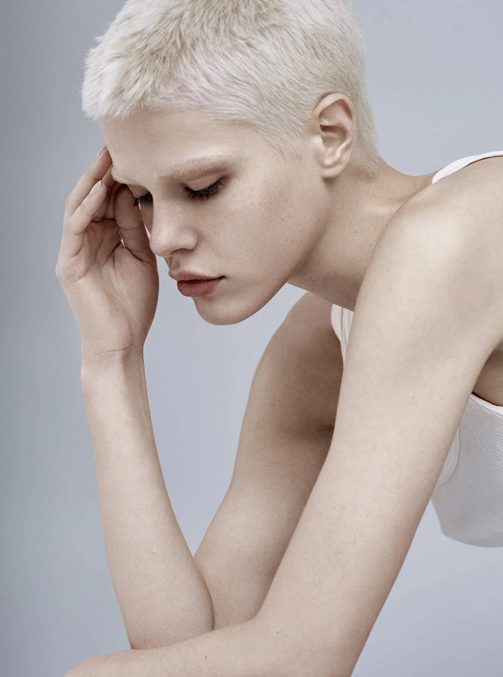 Nahaufnahme eines nachdenklichen Menschen mit kurzen blondem Haar, der seine Stirn berührt, vor einem neutralen grauen Hintergrund.