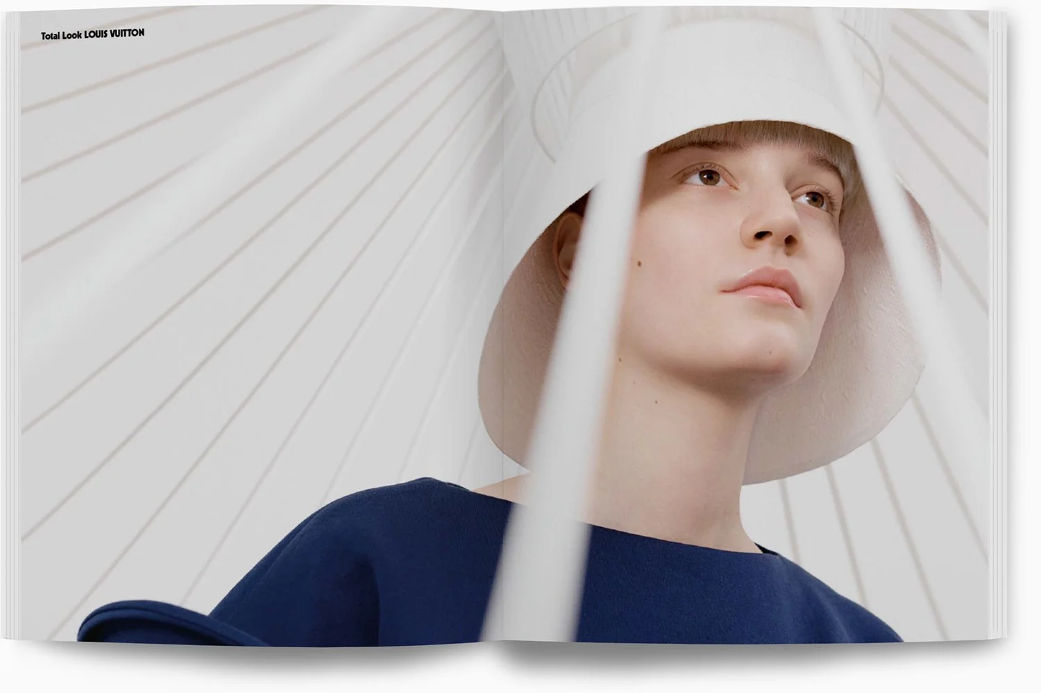 Nahaufnahme eines jungen Mannes mit einem großen weißen Hut, betrachtet nach oben, vor einer weißen, schirmartigen Hintergrundstruktur.