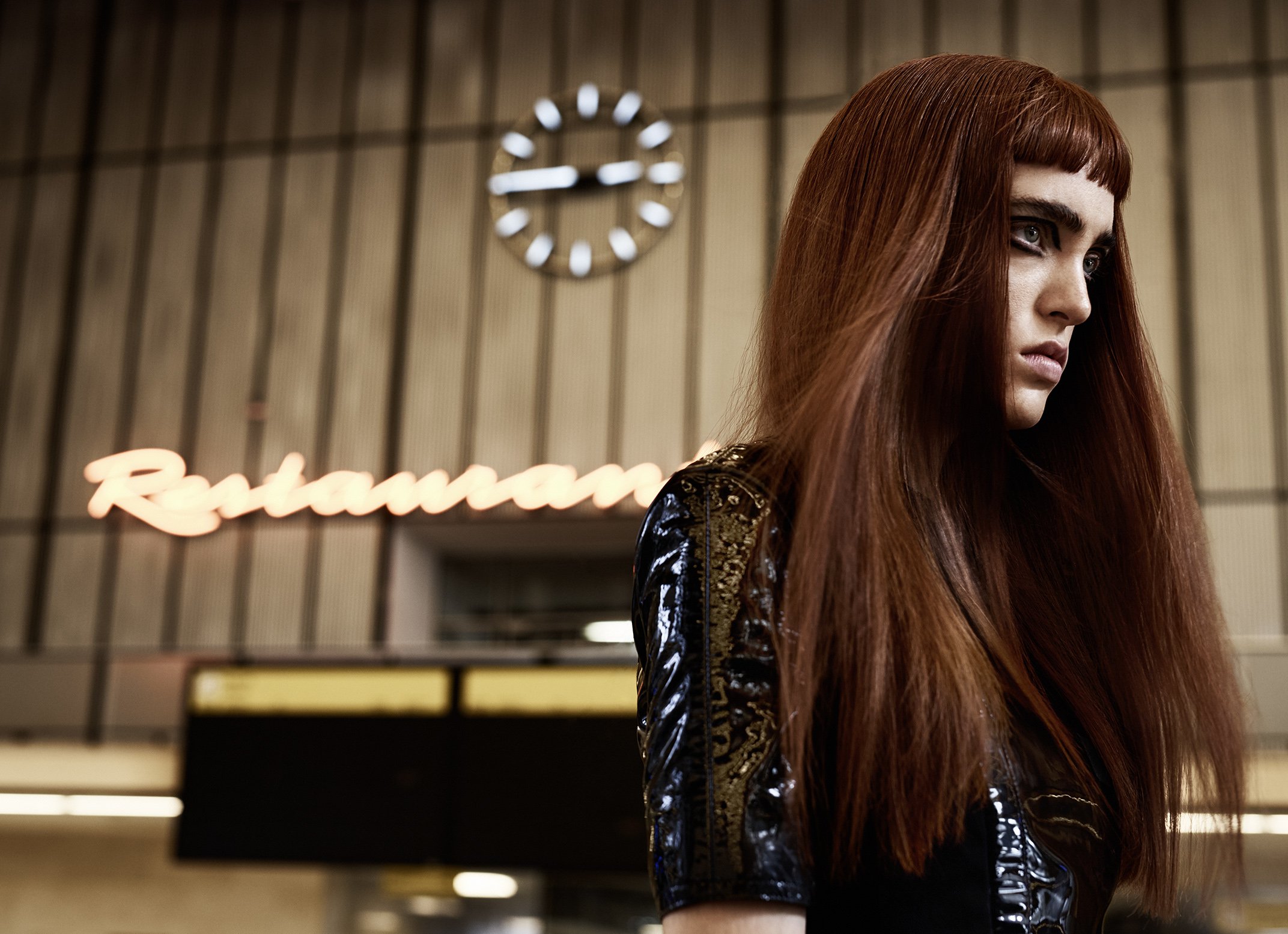 Junge Frau mit roten Haaren und dunklem Make-up in einem Hotel mit einer Uhr und einem Neon-Schild, das 'Restaurant' zeigt.
