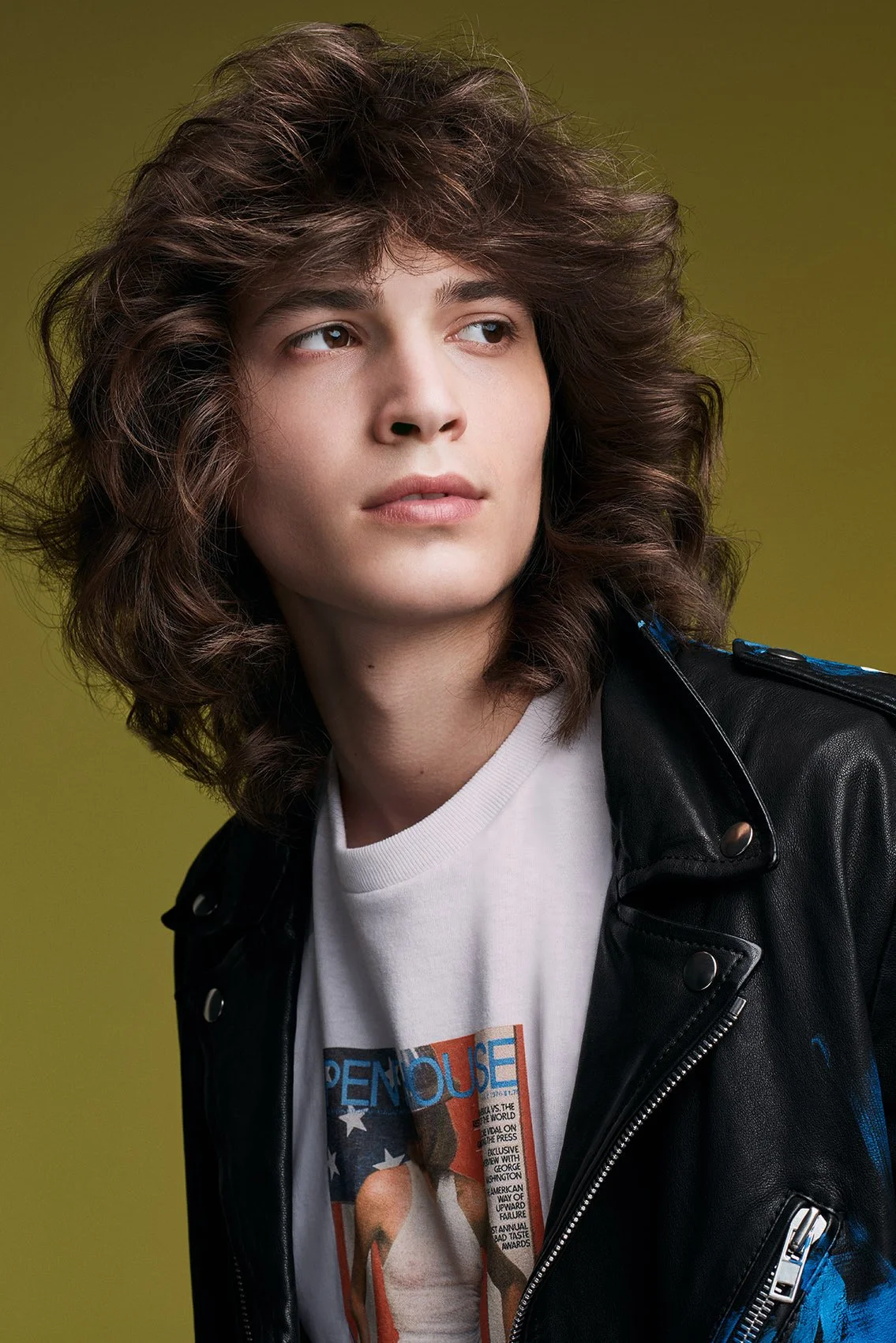 Porträt eines jungen Mannes mit lockigem braunem Haar, trägt eine weiße T-Shirt mit Druck und eine schwarze Lederjacke, vor grünem Hintergrund.