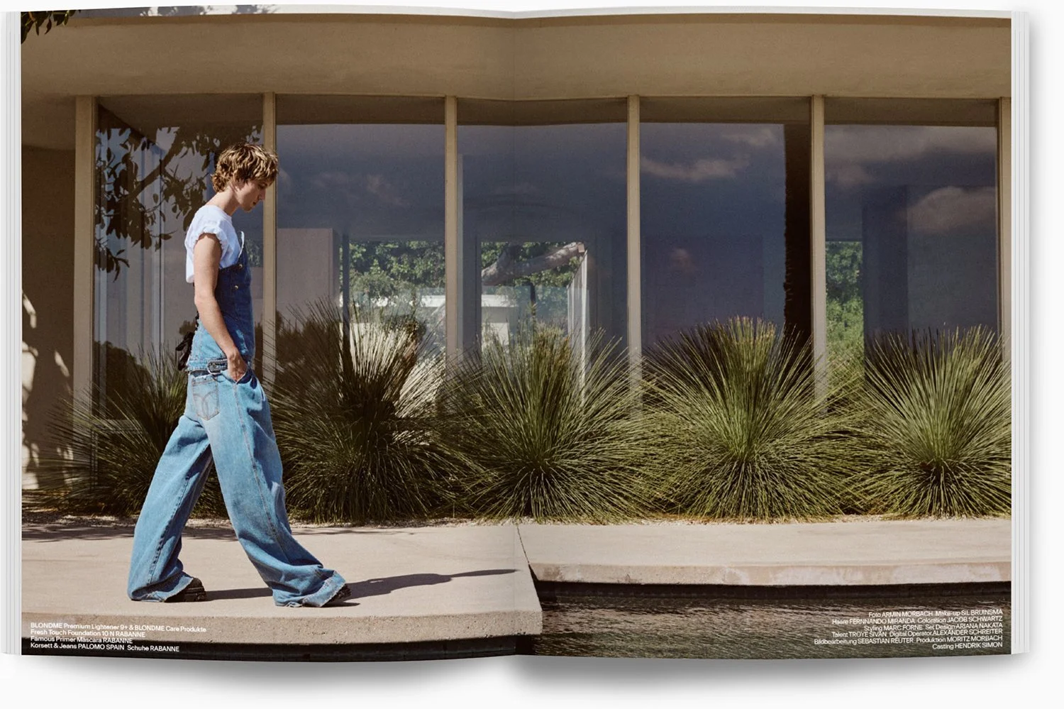 Eine junge Frau in Baggy-Jeans und weißem T-Shirt geht an einer Wasserfläche entlang, im Hintergrund ist ein modernes Haus mit großen Fenstern und Sträuchern zu sehen.