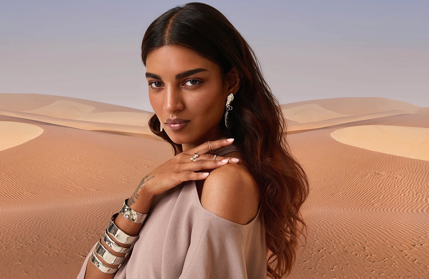A woman with long, wavy brown hair, wearing earrings, rings, bracelets, and a beige top with shoulder cutouts, standing in a desert landscape with sand dunes.