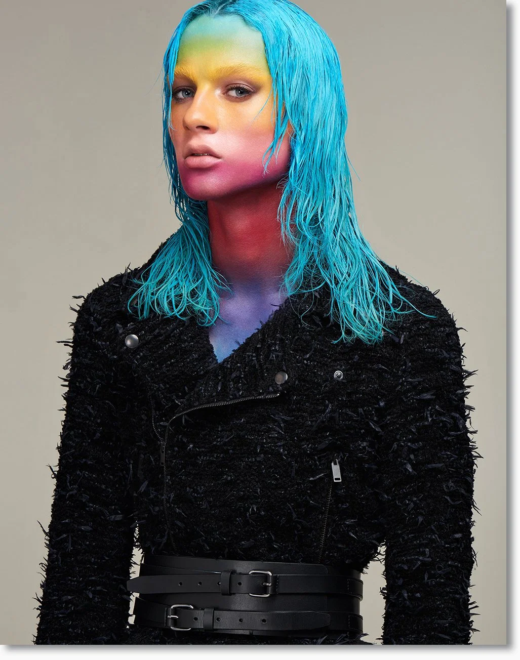 Frau mit buntem Make-up und plätschenden, blauen Haare, trägt schwarze Texturjacke mit Reißverschlüssen und Gürtel, steht vor neutralem Hintergrund.