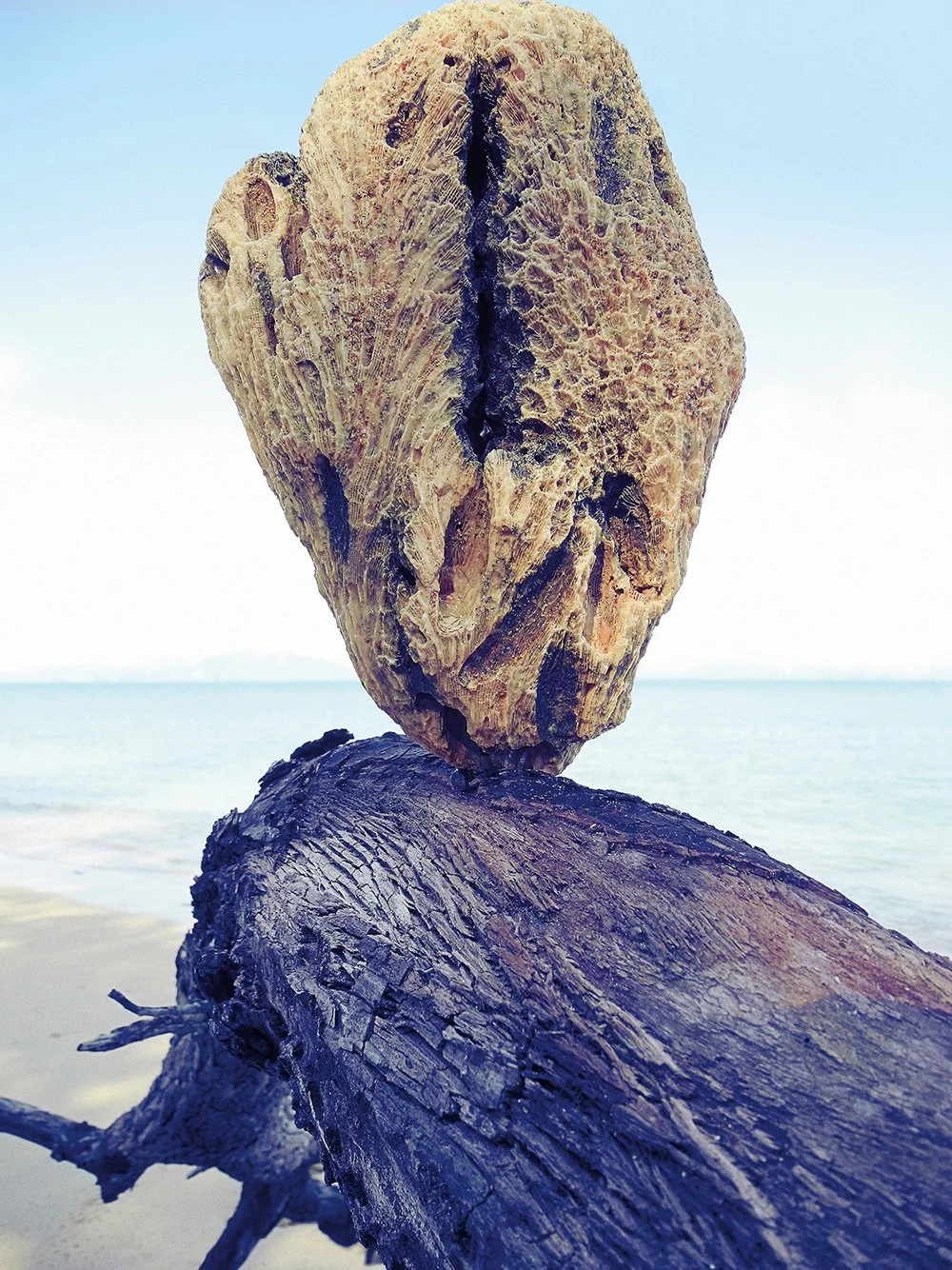 Ein kreatives Kunstwerk aus aneinandergereihten Holzstücken, das an eine abstrakte Person erinnert, am Strand mit Wasser im Hintergrund.