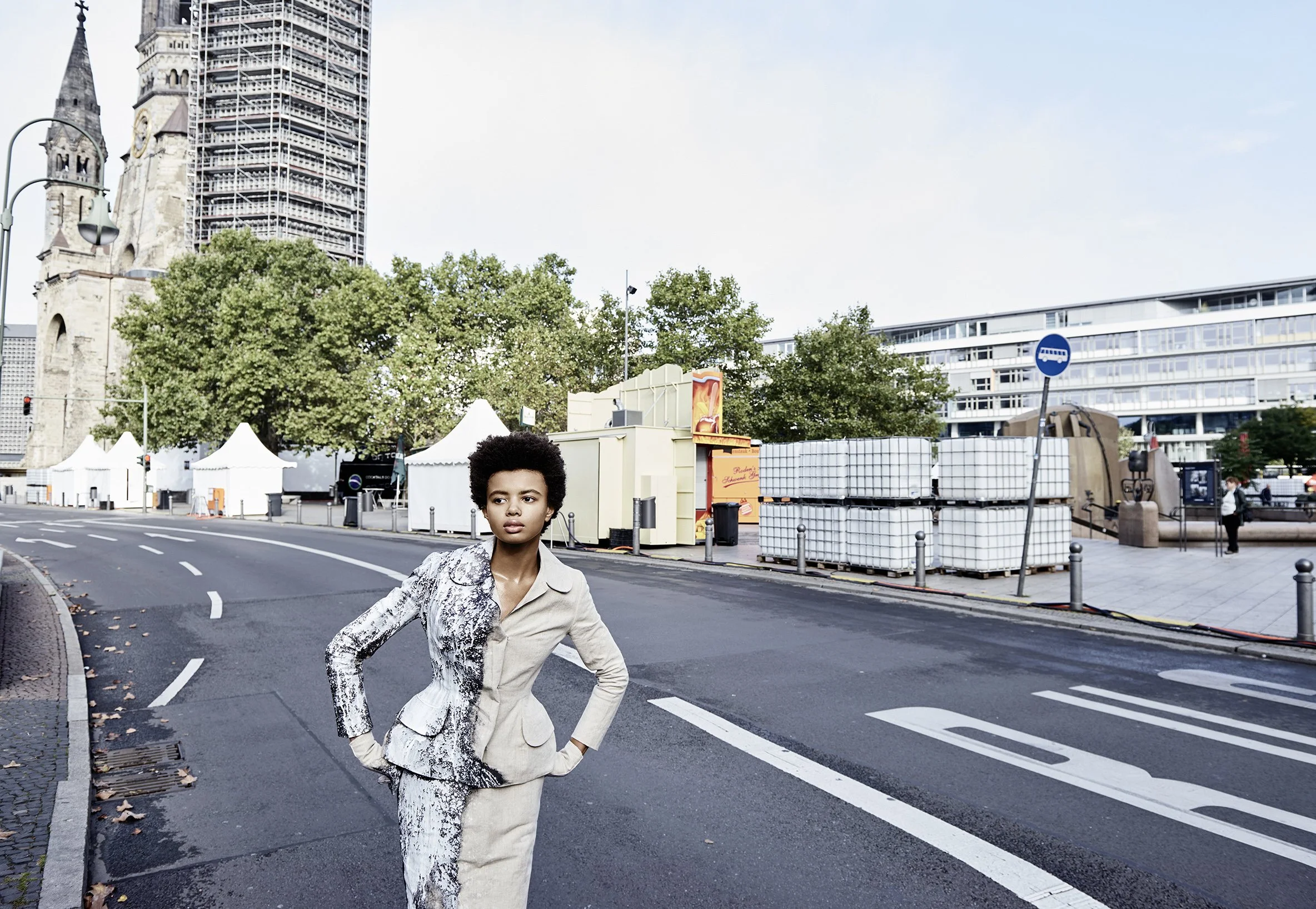 Eine Frau steht auf einer leeren Straße in einer Stadt, im Hintergrund sind Bäume, Bauwerke und Zelte zu sehen.