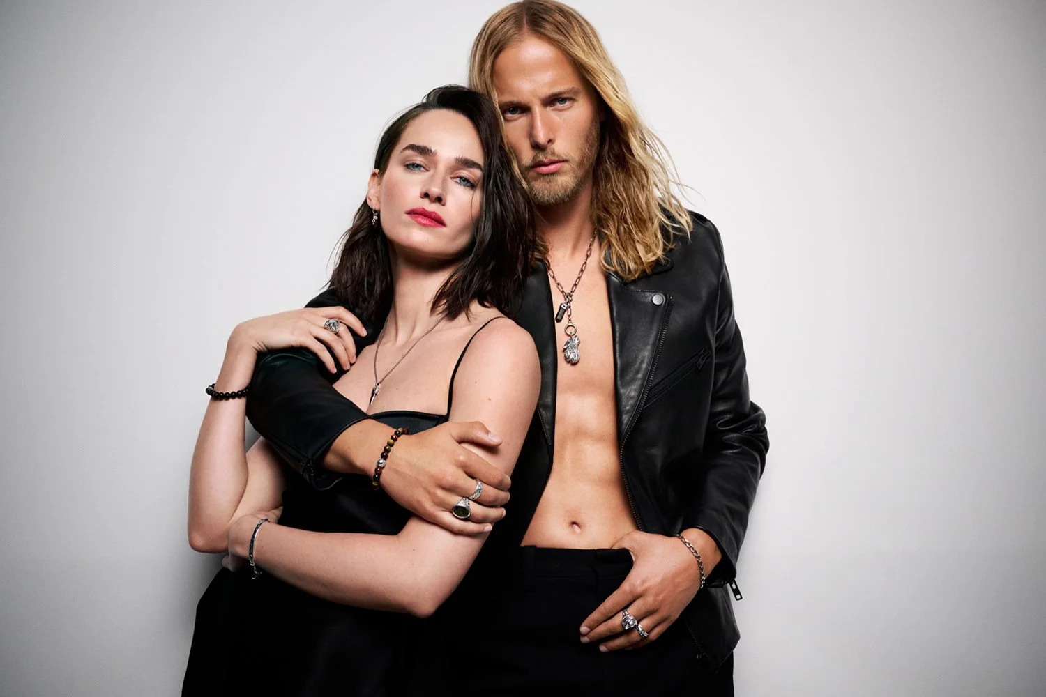 A woman with shoulder-length dark hair and a man with long blonde hair pose together against a plain white background. The woman is wearing a black dress with thin straps and the man is shirtless, wearing a black leather jacket. They are accessorized