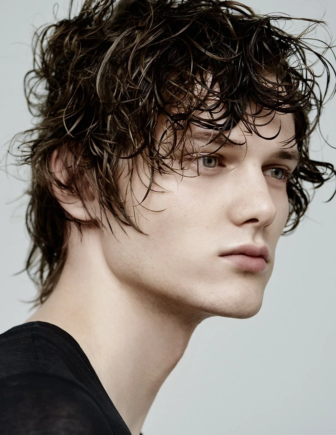 Nahaufnahme eines jungen Mannes mit lockigem braunem Haar und blauen Augen, tragt schwarzen Oberteil.