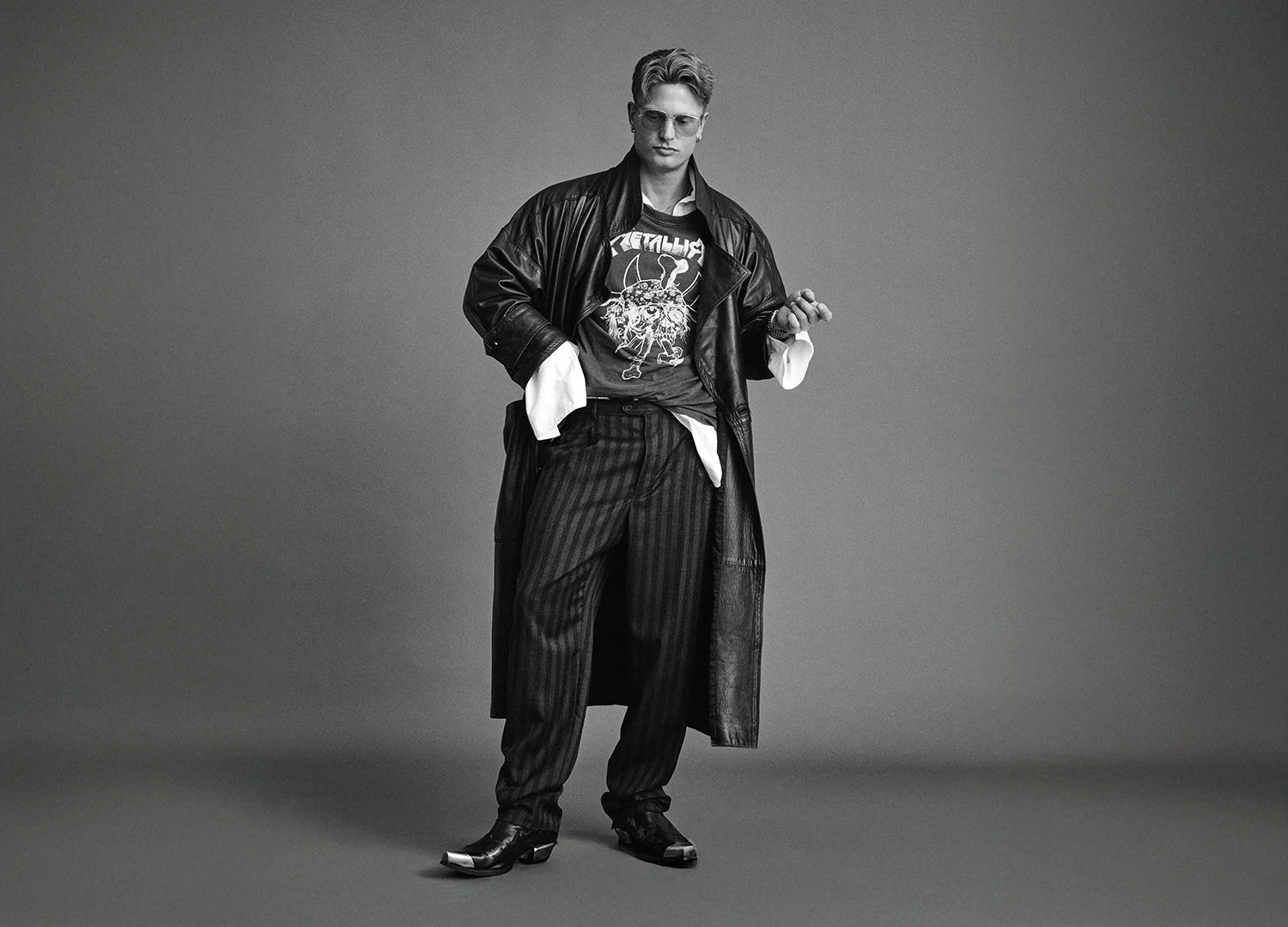 A young man with glasses dressed in a black leather coat, striped trousers, a Metallica t-shirt, and cowboy boots, standing against a plain gray background.