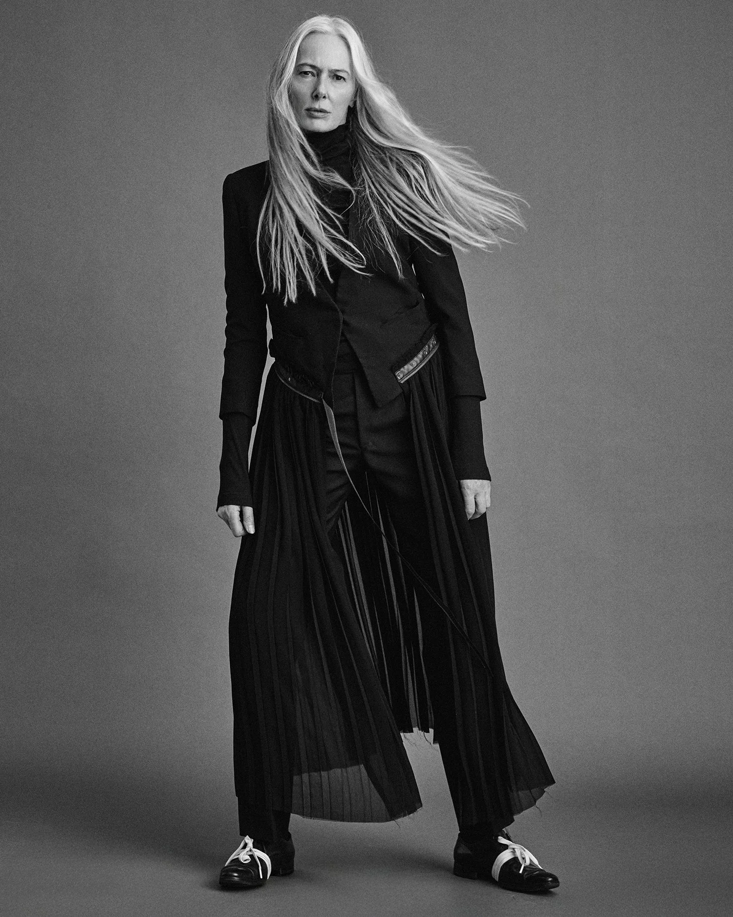 A woman with long, flowing hair in a black and white studio portrait, wearing a dark outfit including a jacket, turtleneck, pleated skirt, and shoes with laces.
