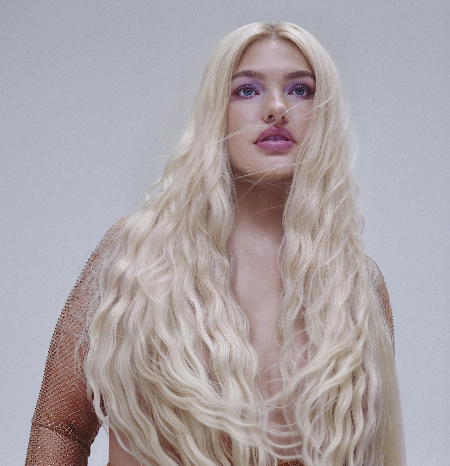 A woman with long, wavy blonde hair and pink lipstick, looking directly at the camera against a plain, light background.