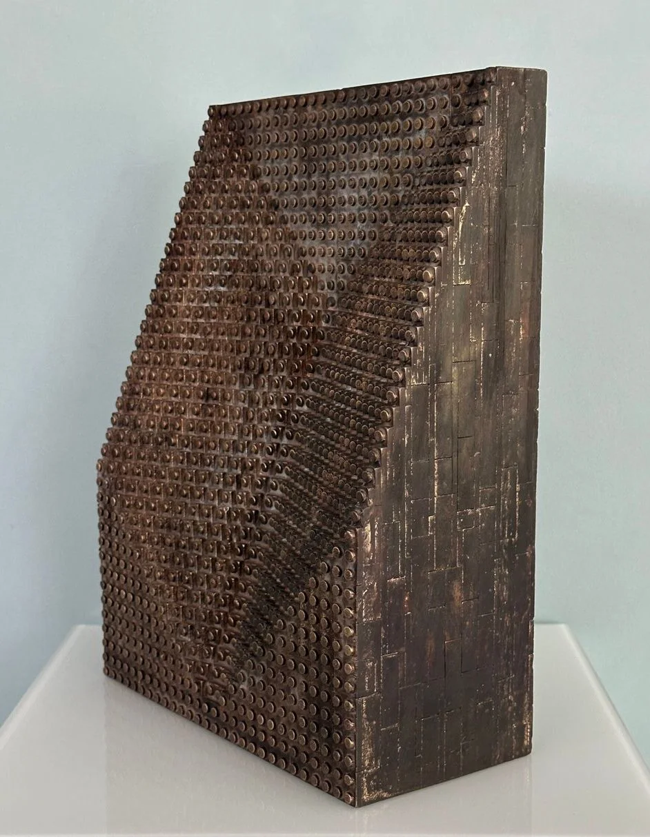 Kunstwerk aus Holz mit hunderten kleinen röhrenförmigen Vertiefungen, die ein menschliches Profil in den Flächen bilden, auf einer weißen Plattform vor hellblauer Wand.