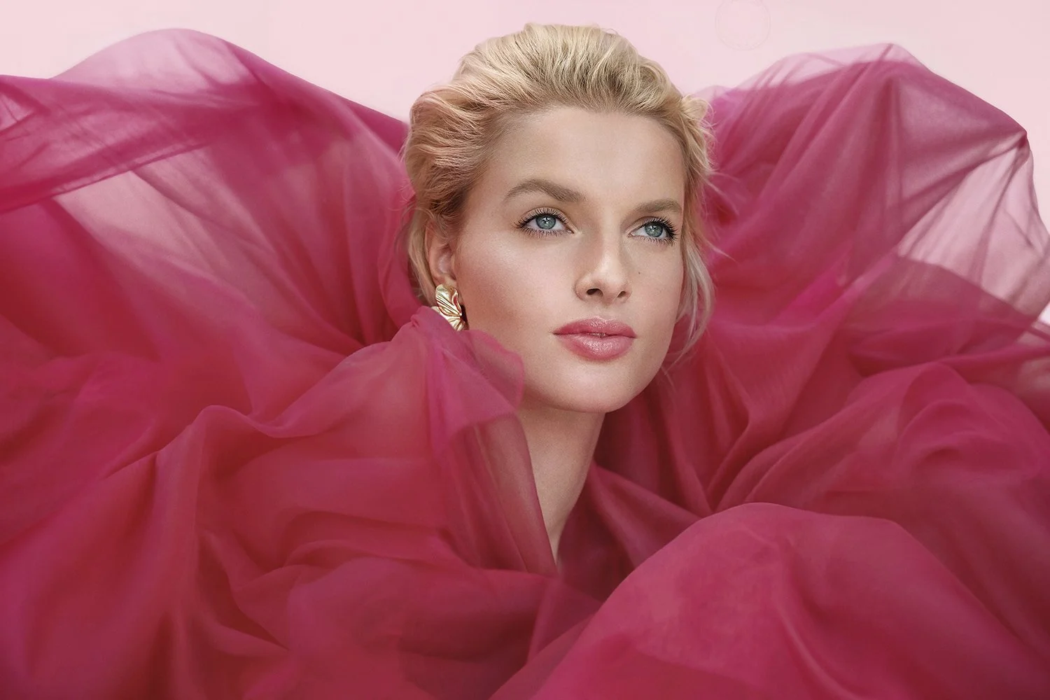 A woman with blonde hair and blue eyes wearing gold earrings, surrounded by flowing pink fabric, against a pink background.