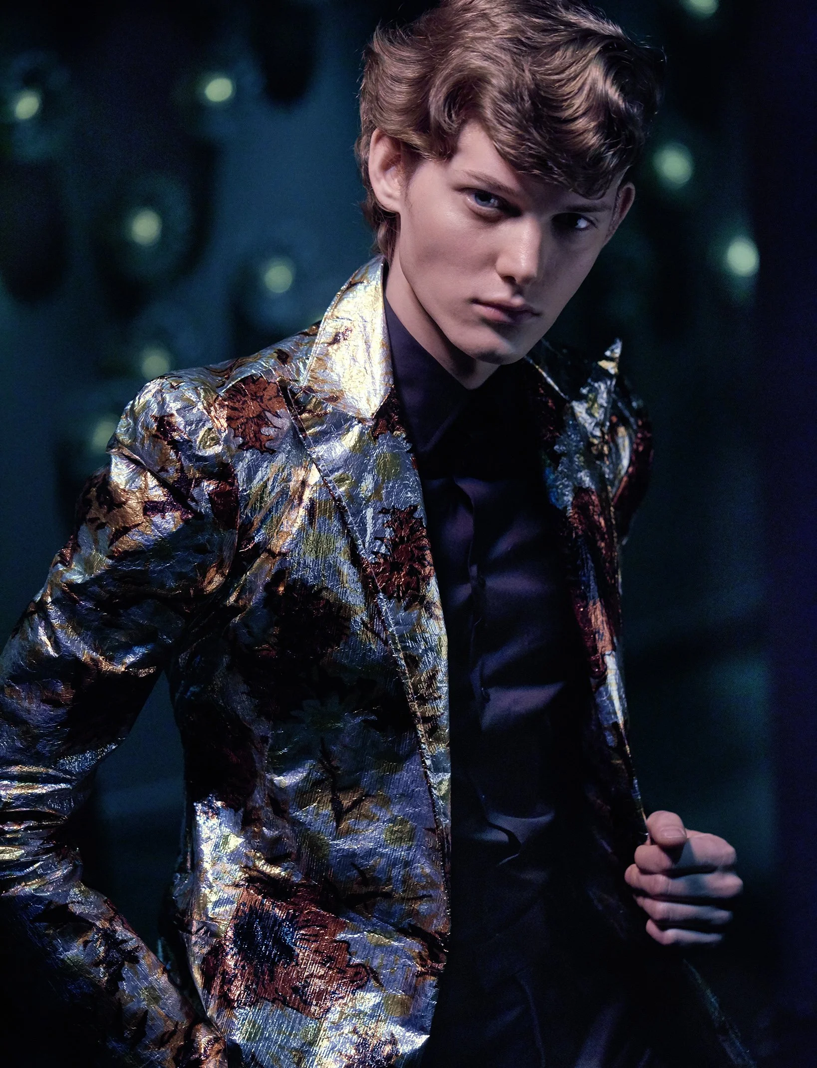 Junge Mann in einem schimmernden, floralen Jackett, schwarzes Hemd, dunklem Hintergrund mit grünen Lichtern.