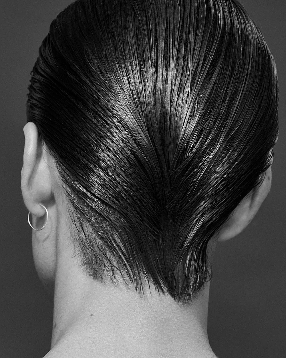 Schwarzes, nasses Haar vom Hinterkopf sichtbar, mit Ohr und Ohrring auf der linken Seite, in schwarz-weißer Aufnahme.