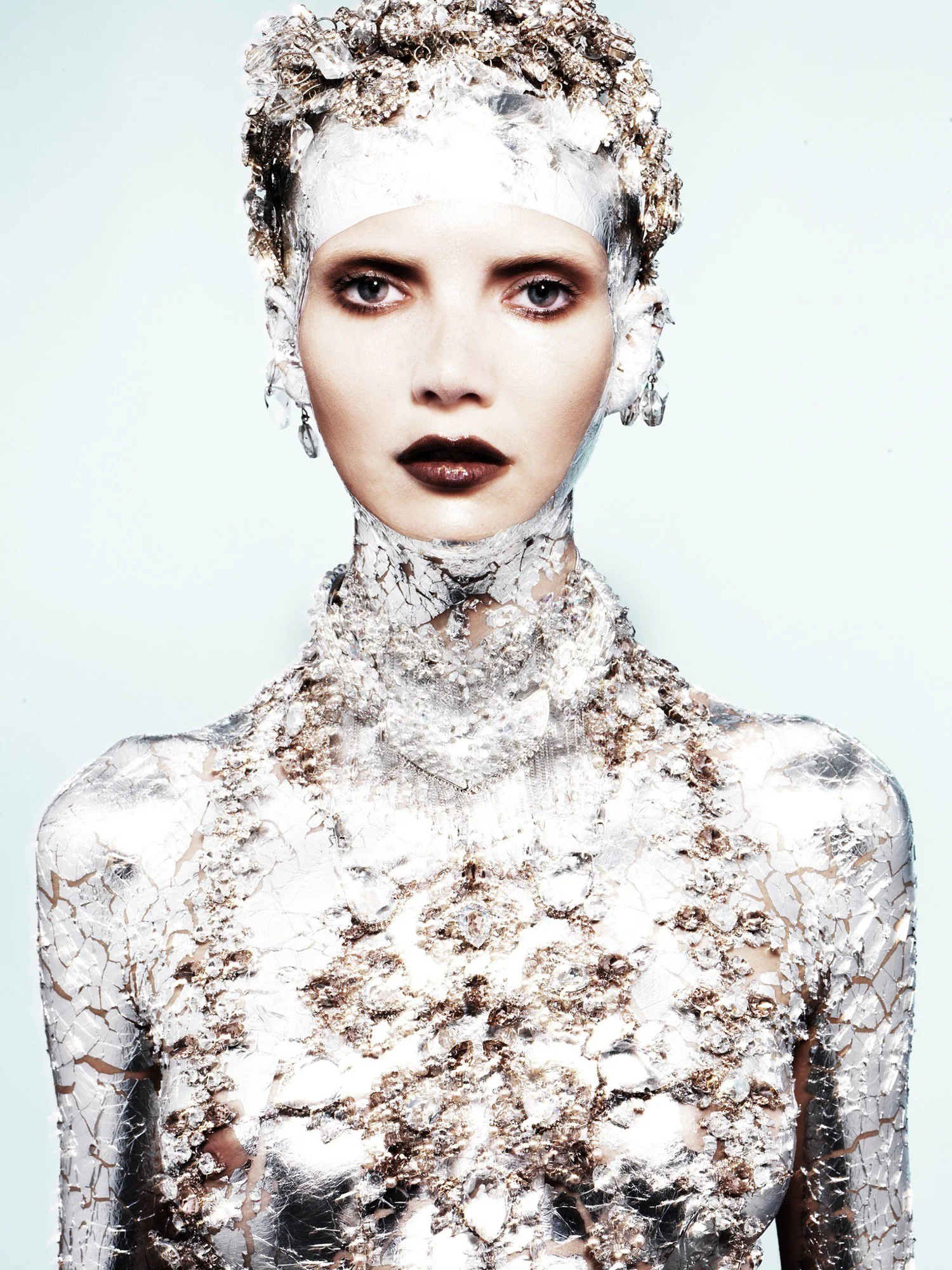 A woman with pale skin and dark makeup, wearing a headpiece and body covered in cracked, reflective silver material.