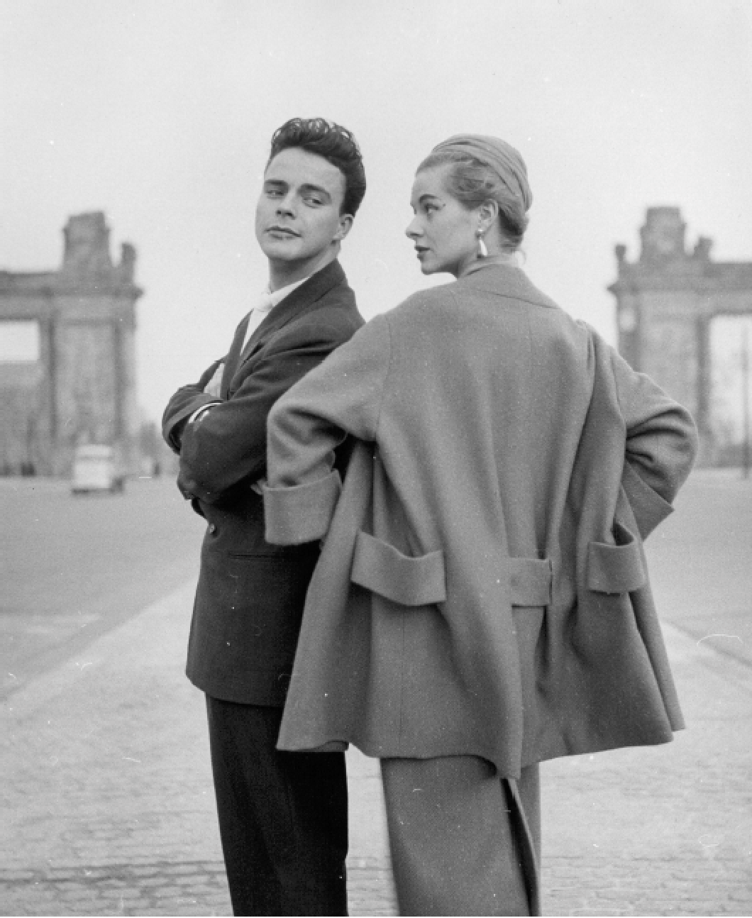 Ein schwarzes und weißen Fotografie von einem Mann und einer Frau, die sich auf einer Straße in einer Stadt befinden. Der Mann trägt einen Anzug, während die Frau einen Mantel trägt. Im Hintergrund sind Gebäude sichtbar.