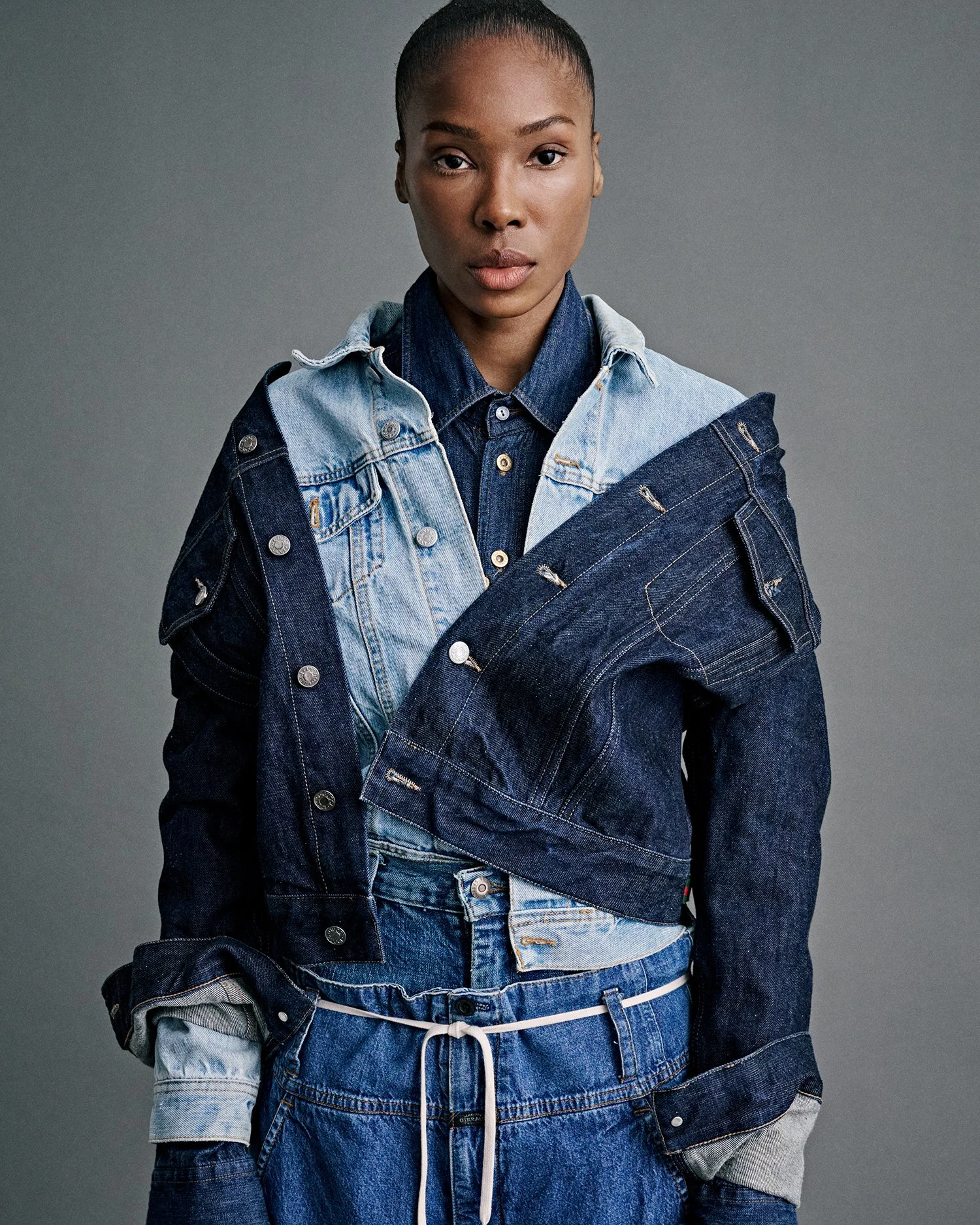 A woman wearing a layered denim jacket over a denim shirt and jeans, standing against a plain gray background.