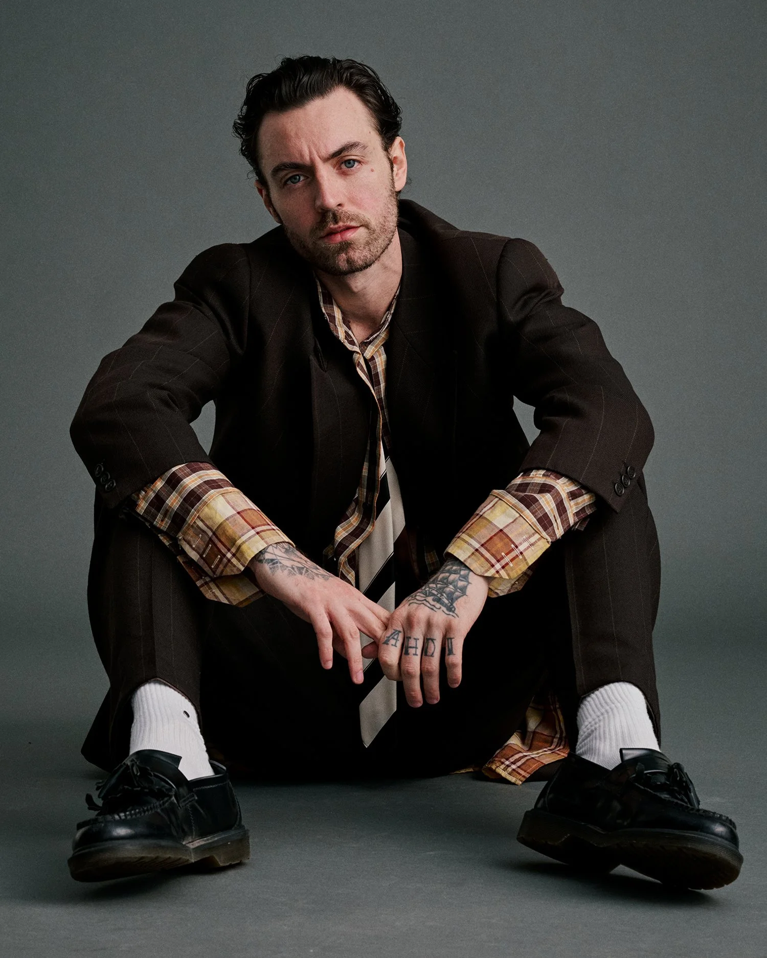 A man with dark hair, tattoos, and a beard sitting on the floor against a gray background, wearing a black blazer, plaid shirt, tie, black pants, white socks, and black shoes.