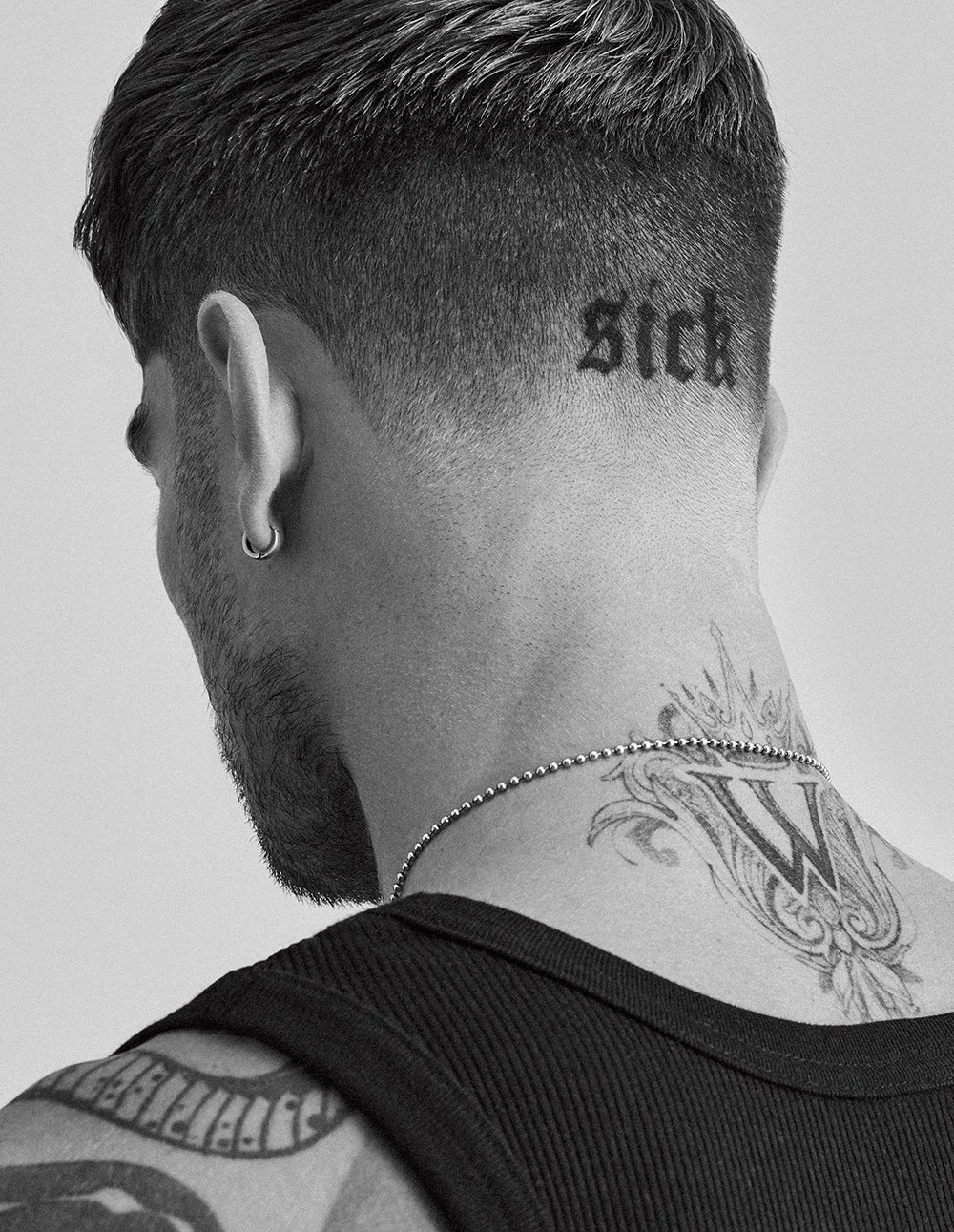 Ein Mann mit kurzen, dunklen Haaren und Tattoos am Nacken, trägt ein Ohrring und eine Halskette, gesehen von hinten in Schwarz-Weiß.