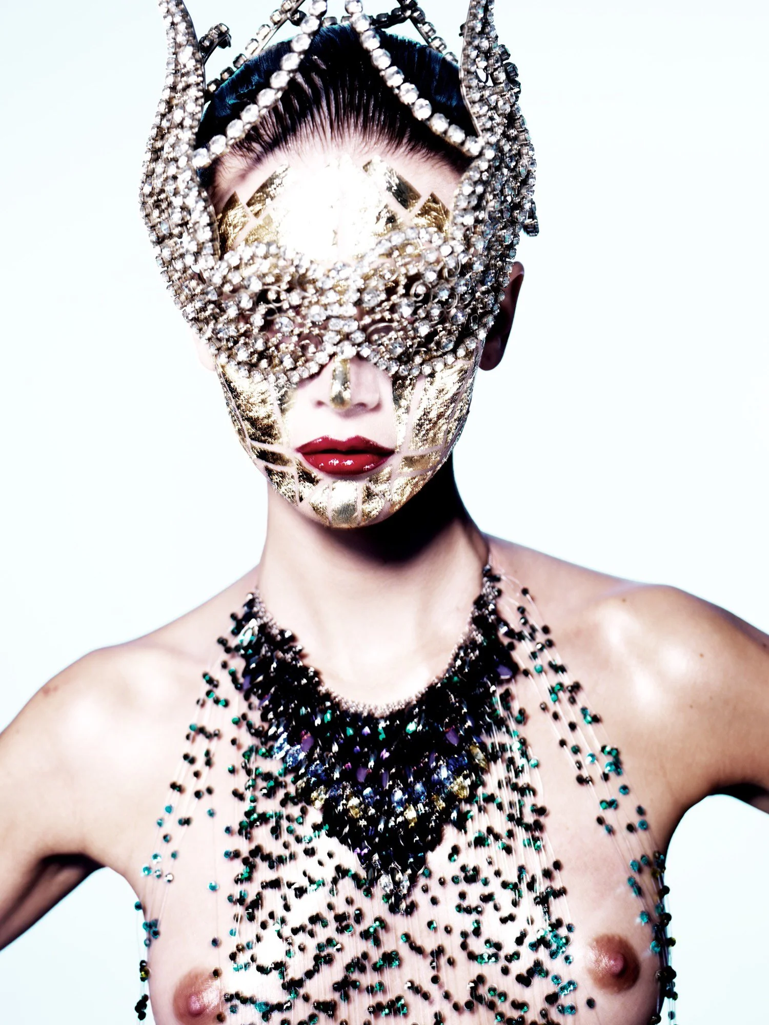 A person wearing an ornate mask adorned with rhinestones and gold details, covering the upper face. They have bold red lipstick, dark hair styled back, and multiple layered necklaces with beads and stones. The person is topless, with the necklaces dr