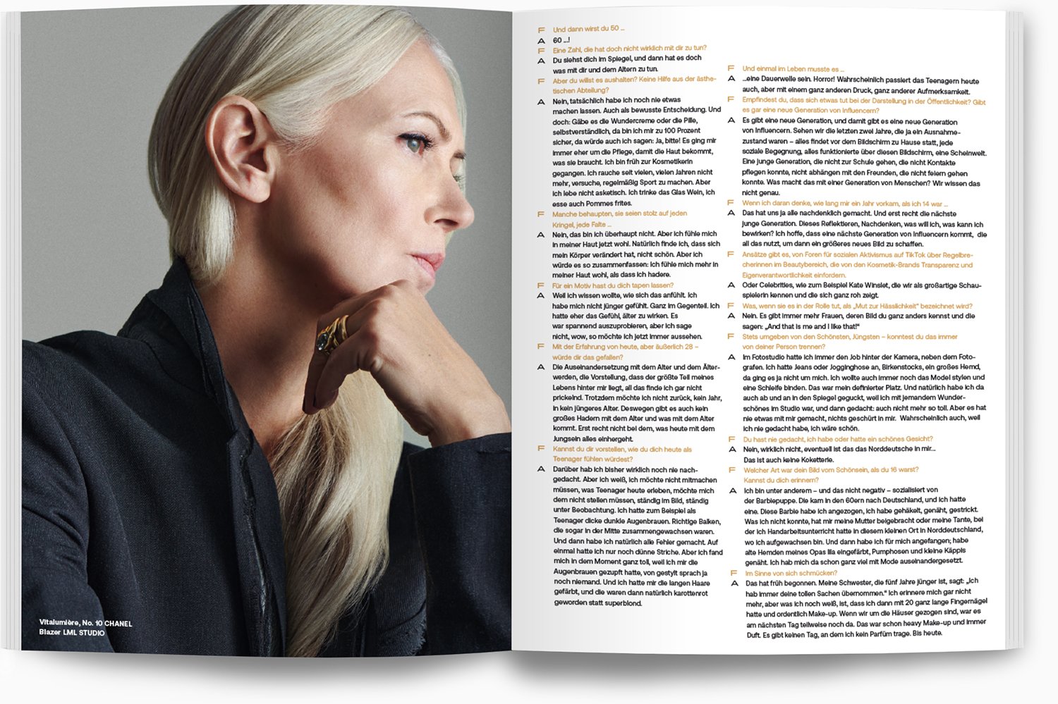 Eine ältere Frau mit blondem, glatt geführtem Haar, trägt einen schwarzen Blazer und eine große goldene Halskette. Sie sitzt nachdenklich und stützt ihren Kopf auf ihre Hand, mit einem goldfarbenen Ring an ihrem Finger.