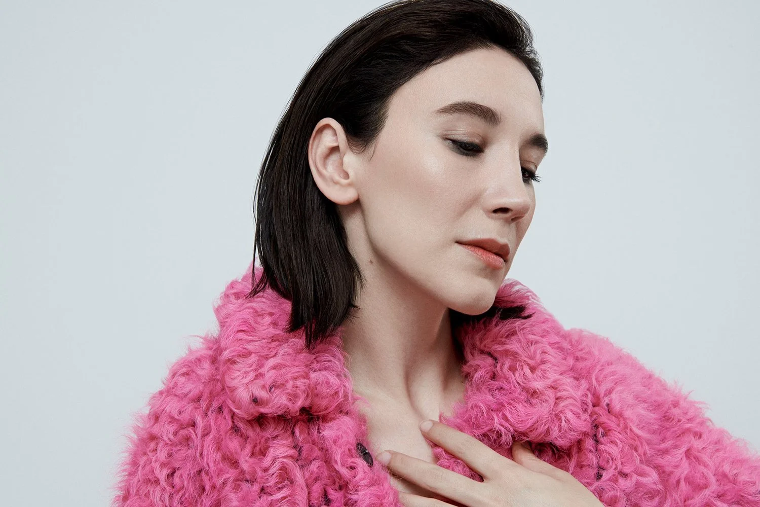 A woman with dark hair wearing a pink fuzzy coat, looking downward with one hand over her chest.